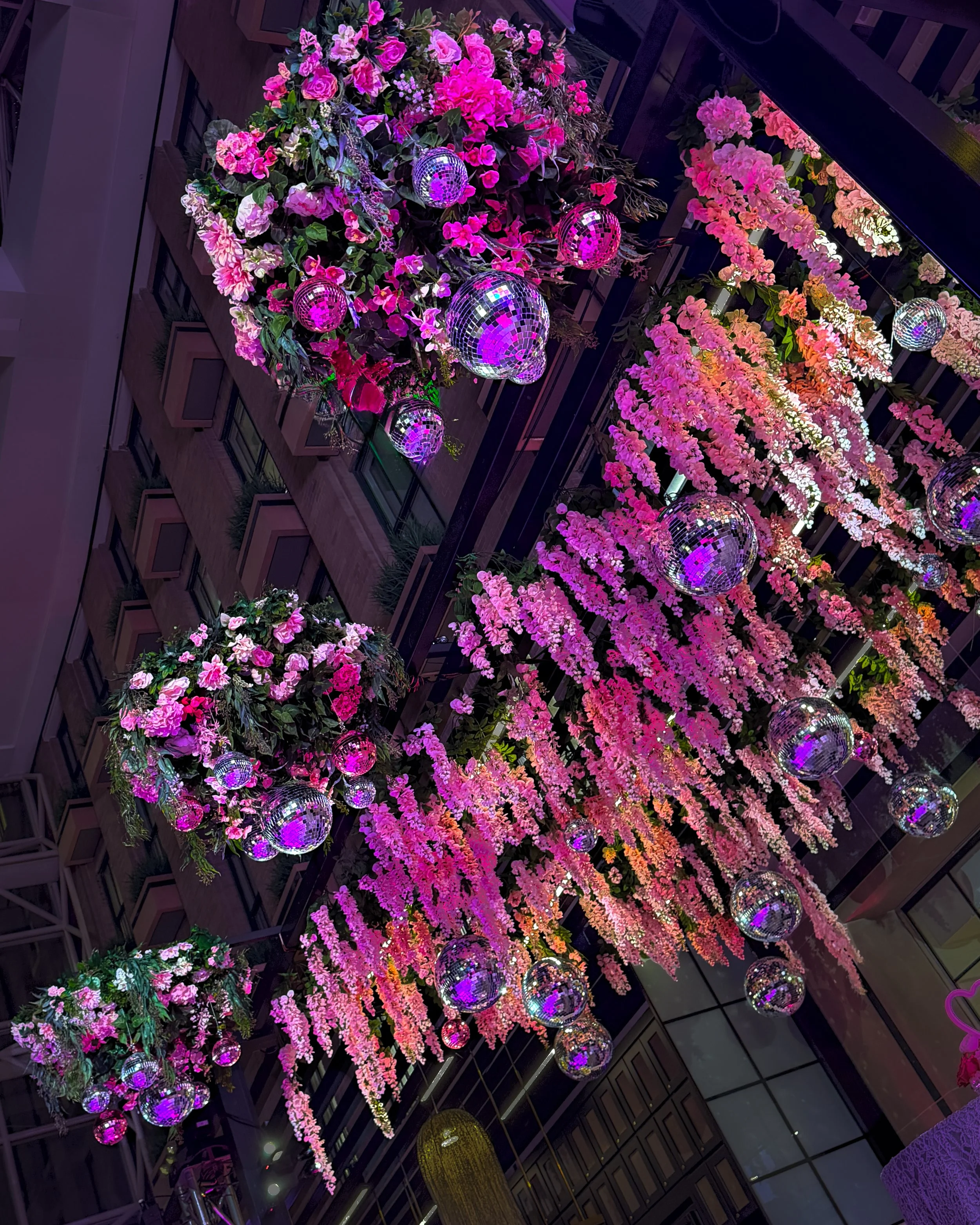 Decorative hanging floral arrangement with pink and purple flowers and disco balls reflecting purple light, hanging from a ceiling with dark support beams and glass windows.