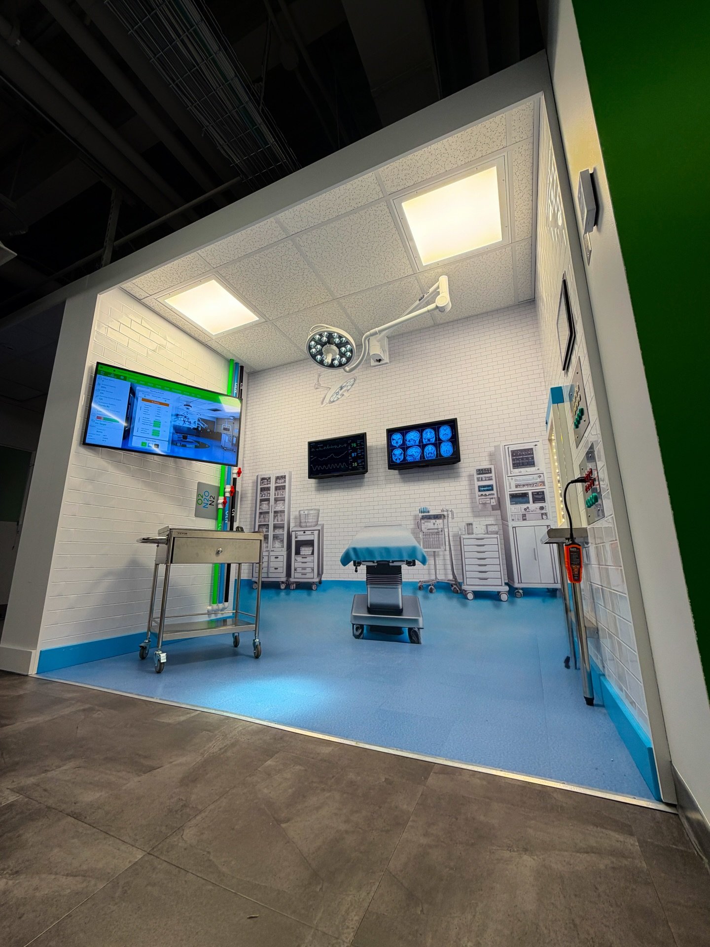 A medical simulation room with hospital equipment, monitors displaying brain scans, and surgical tools, set against white brick walls with bright ceiling lights.