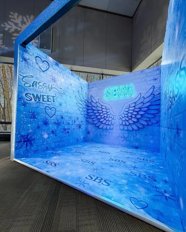 Photo of a decorated winter-themed photo booth with blue snowflake designs and neon signs that say "Sienna" and "Sassy and Sweet."