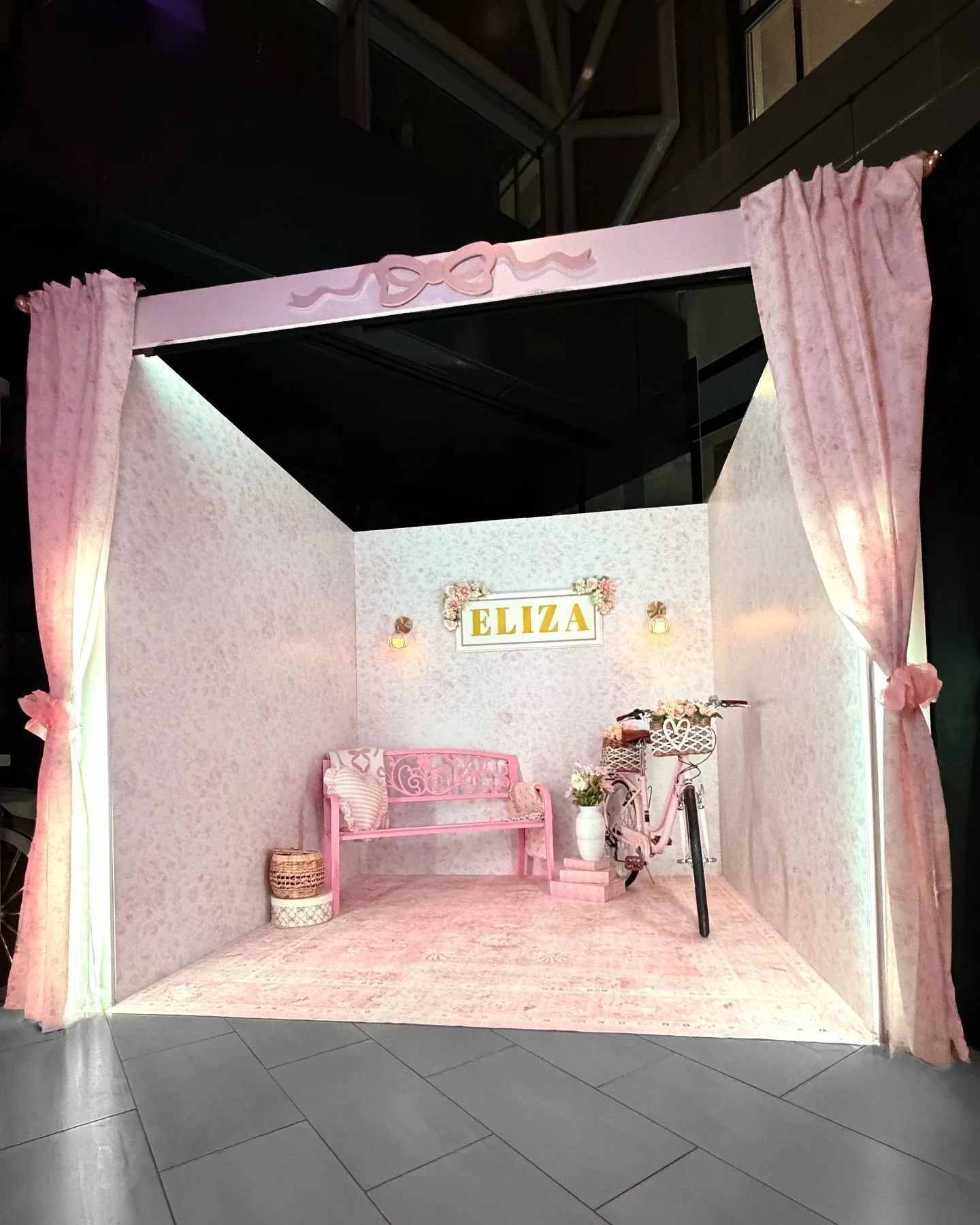A decorated photo booth setup with pink curtains, a pink bench, a pink bicycle with a basket, a white vase with flowers, and a sign with the name 'Eliza' above it, all in a pink-themed background.