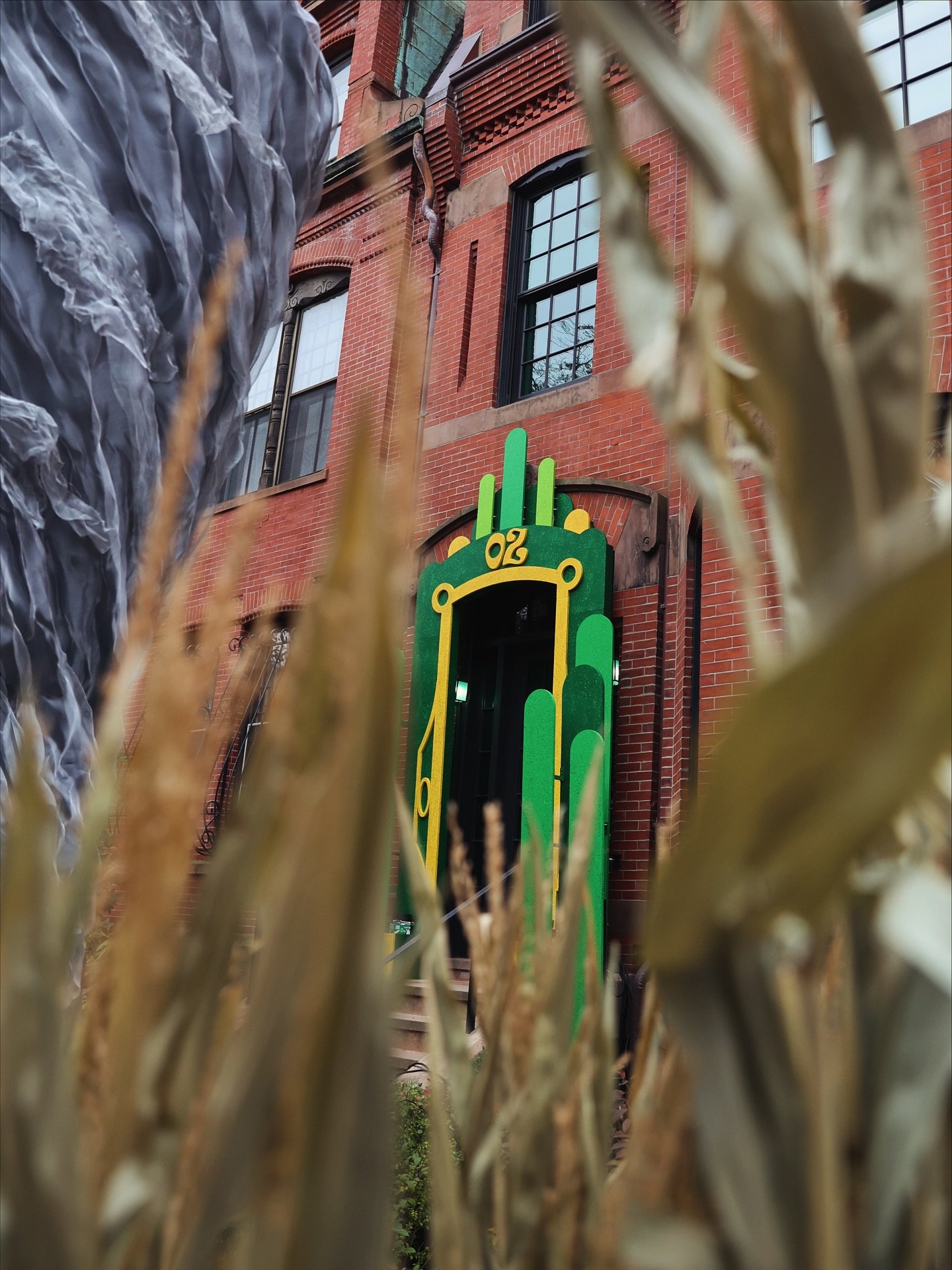 View of a red brick building through a metal fence or barrier with dry vegetation in the foreground. The building has black-framed windows and decorative architectural details. There is green and yellow signage with the number 02.