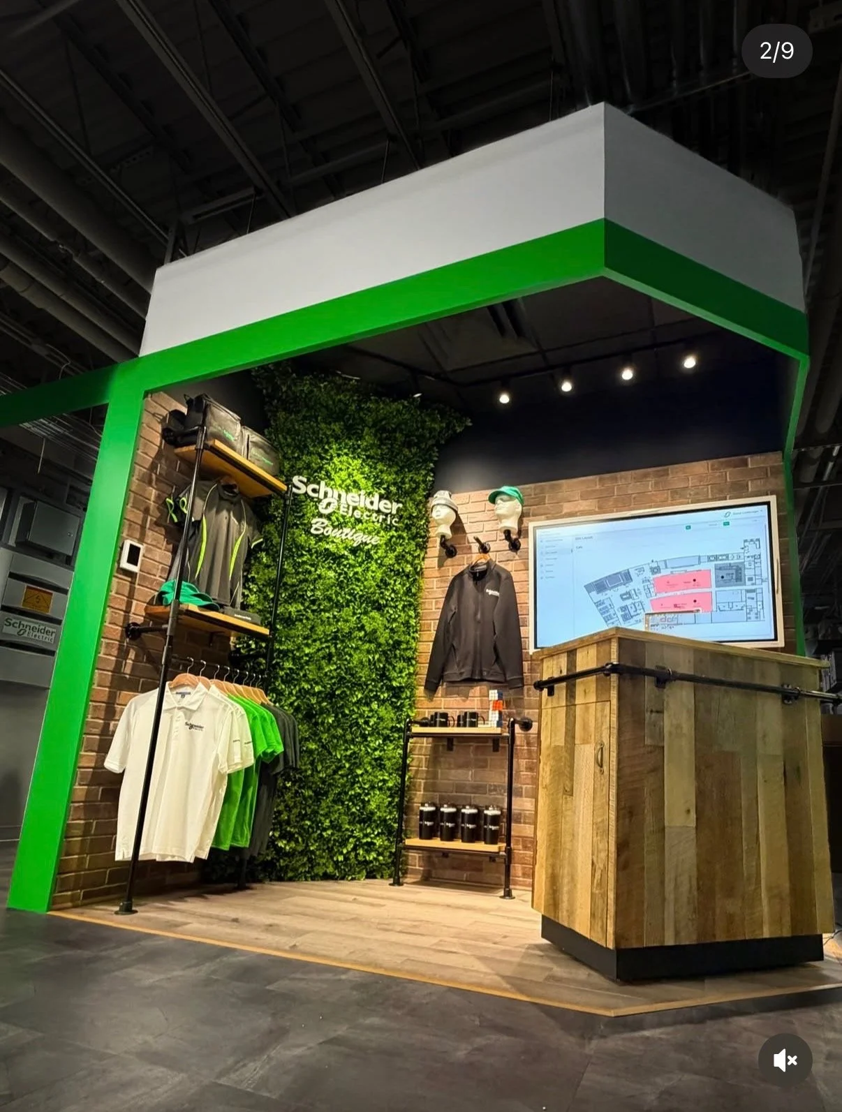 Booth display for Schneider Electric with branded apparel, hats, and accessories, featuring a green and white color scheme, a digital floor plan on a screen, and a backdrop with greenery and the Schneider Electric logo.