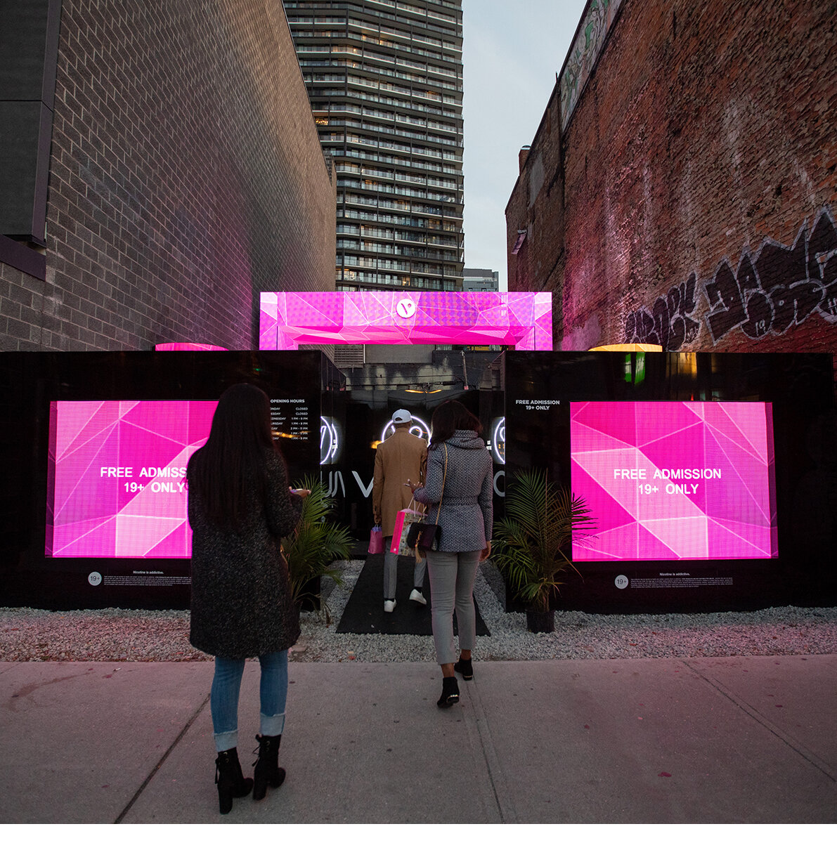 3 people all dressed for cold weather entering into the Vype brand activation in downtown Toronto.  2 LED screens on either side of them and big neon pink LED arch in the background.