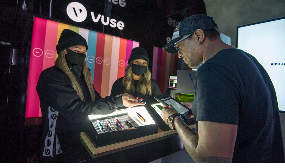 1 man wearing a black t-shirt and black hat, signing up during the Vuse brand activation sampling program. 2 female brand ambassadors assisting him with the signup process.