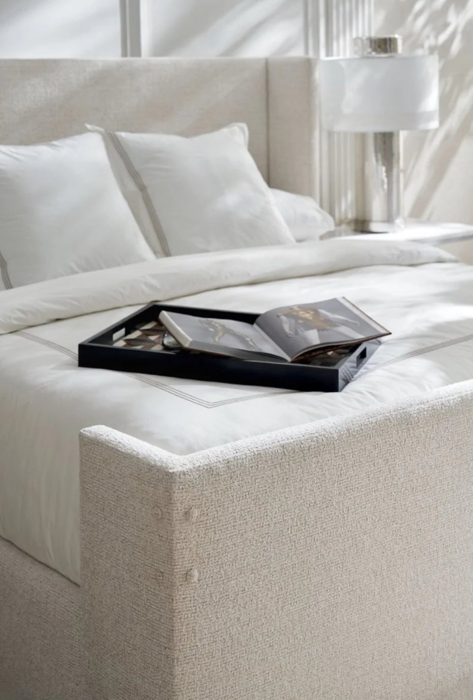 Elegant bedroom featuring a light upholstered bed with crisp white bedding, styled with a black tray holding an open book. A modern table lamp on the bedside table adds soft lighting, creating a peaceful and refined atmosphere.