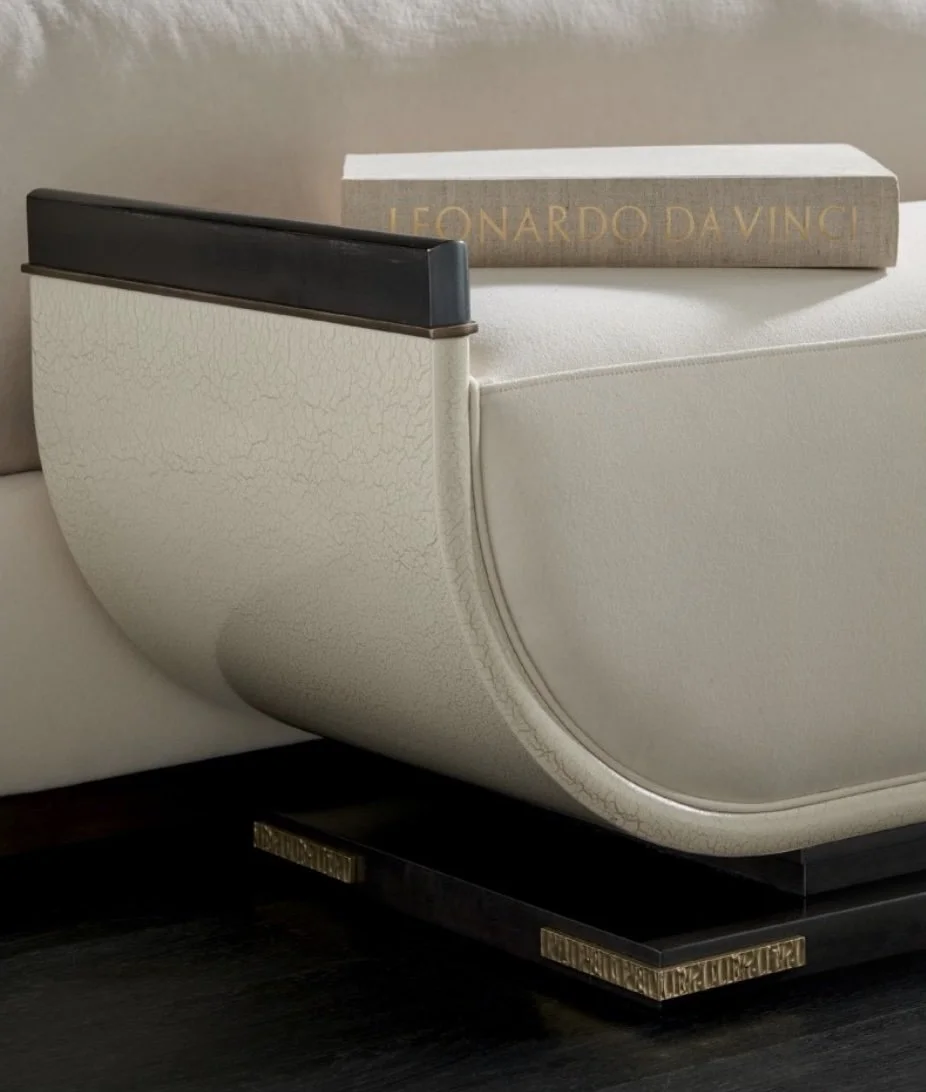 Close-up of a luxurious chair with cream textured upholstery and a dark wooden armrest. A hardcover book titled 'Leonardo Da Vinci' rests on the arm, adding to the refined and elegant design, with intricate gold accents on the base.