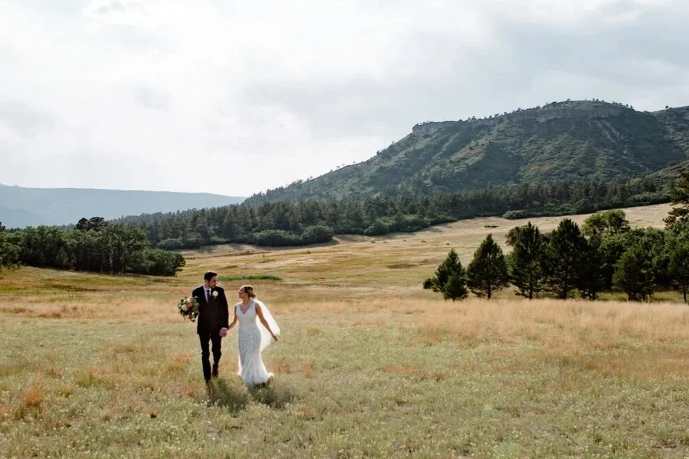 A bride and groom walking hand in hand through a field at Dawson Butte in Colorado before their backyard wedding.