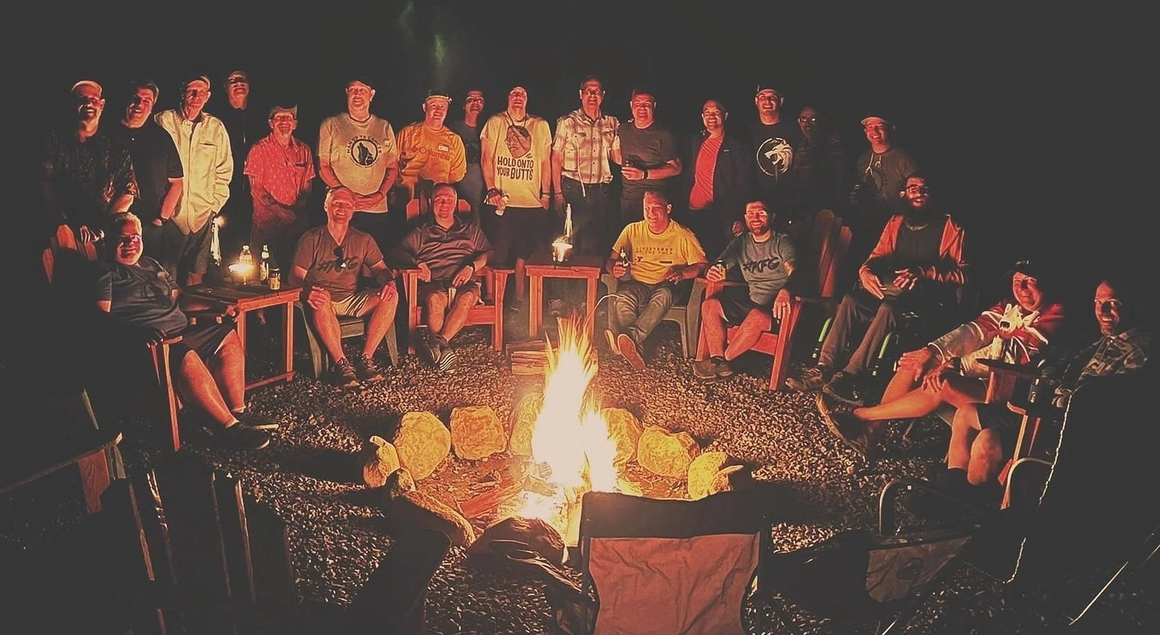 A group of people sitting and standing around a campfire outside at night, with some holding drinks and smiling at the camera.