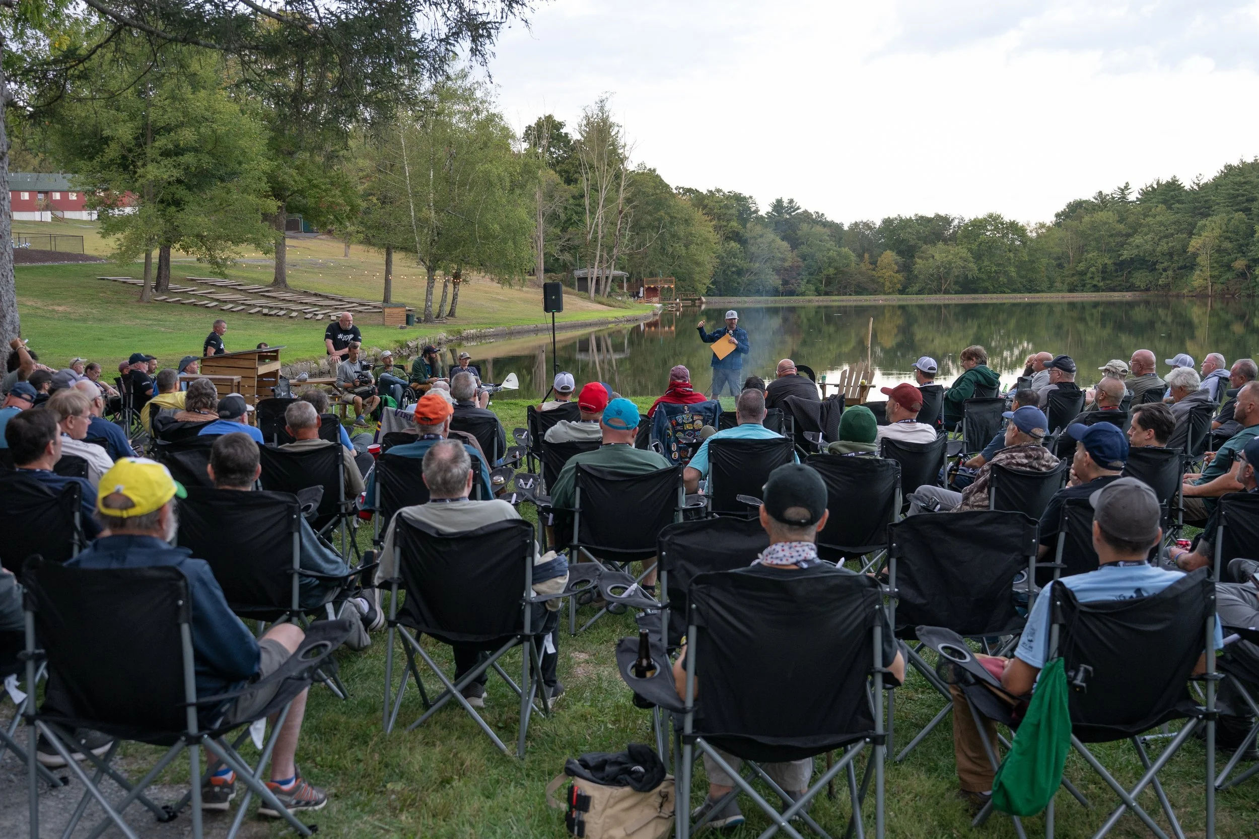 A large outdoor gathering by a lake with many people seated in lawn chairs, listening to a speaker standing with a microphone near the water. The scene is surrounded by trees and grassy areas, with additional seating and pathways visible in the background.