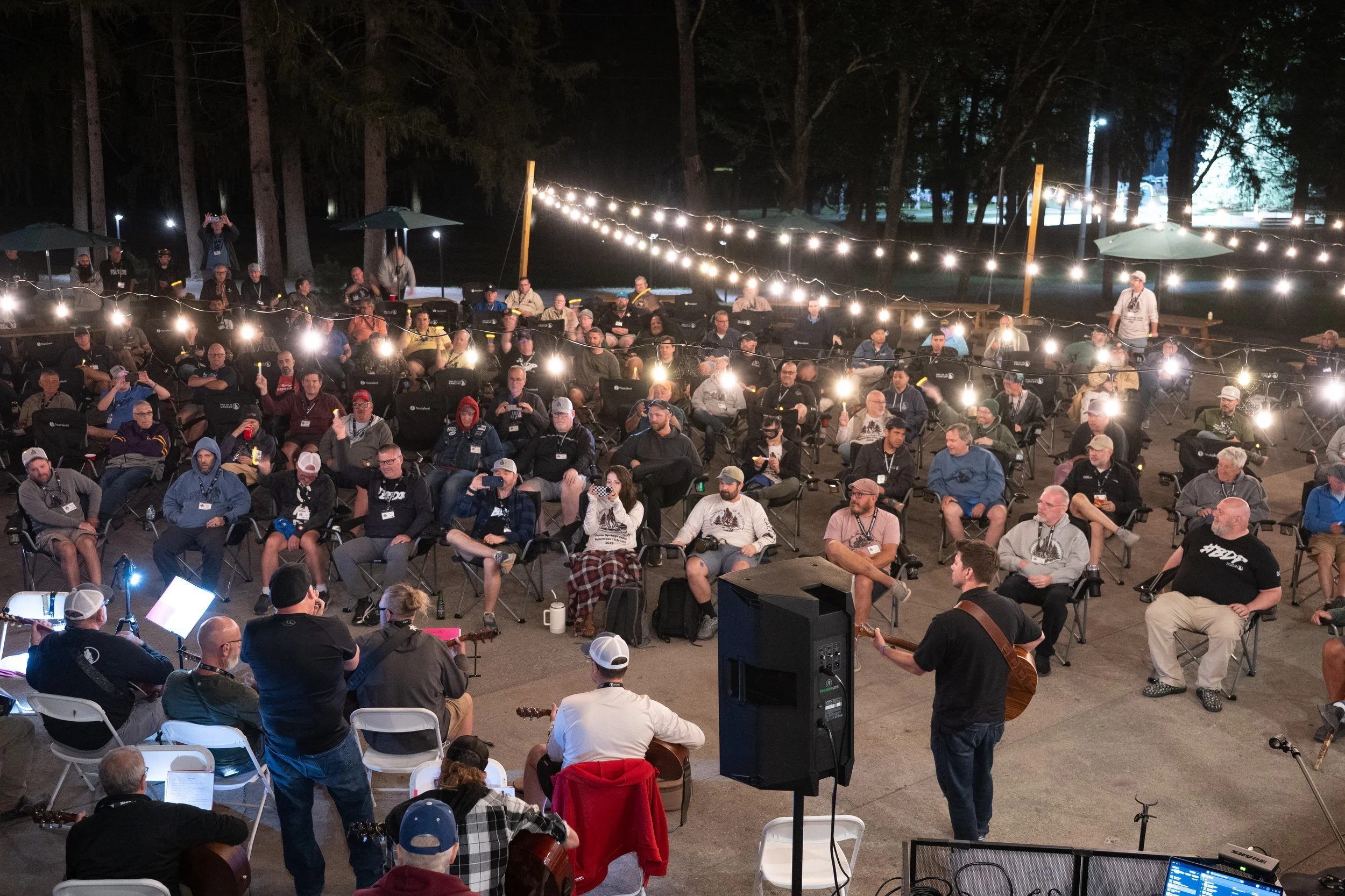Outdoor nighttime gathering of people seated in folding chairs, illuminated by string lights, with some musicians playing guitars at the front of the audience.