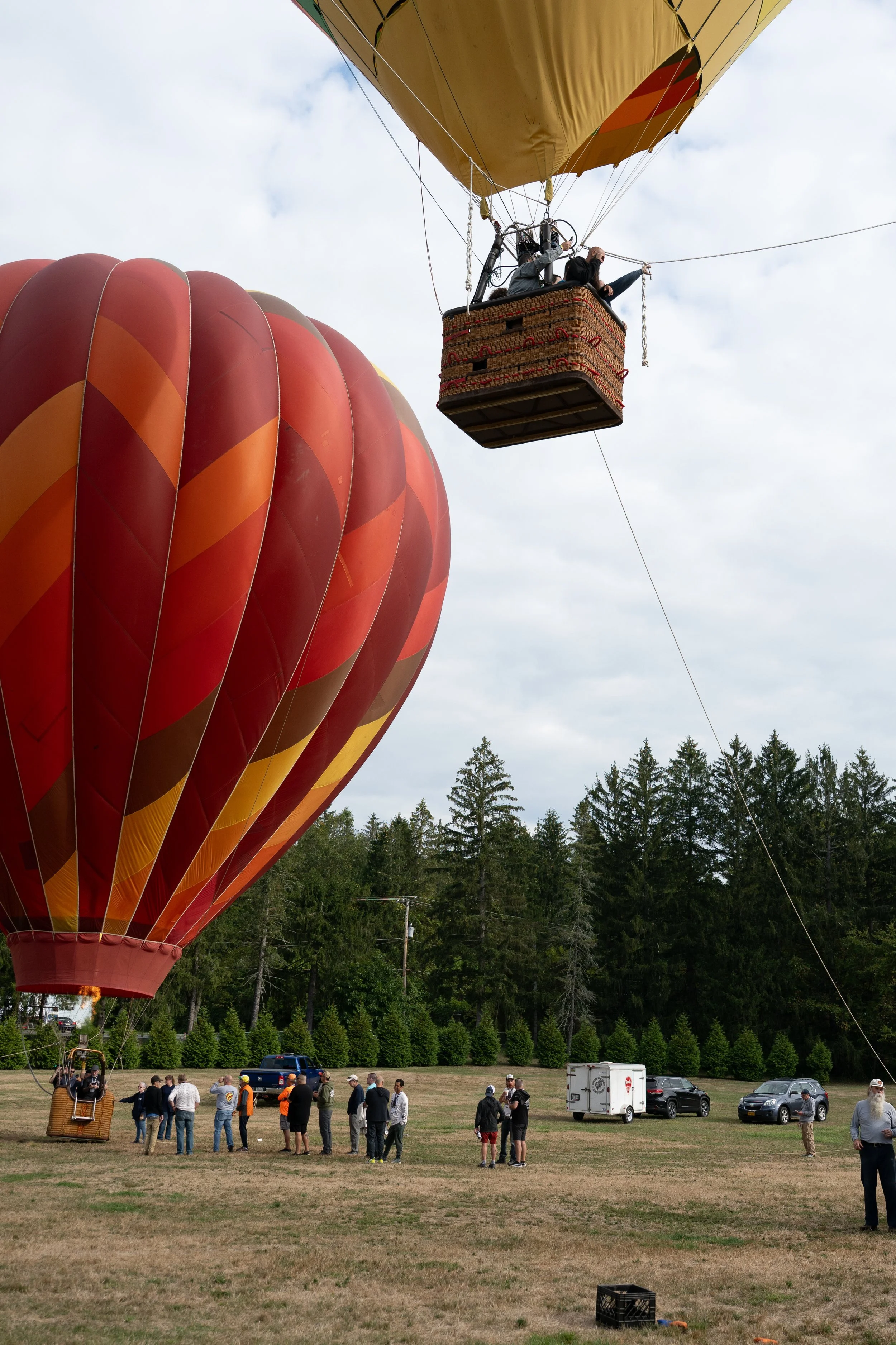 A hot air balloon with an orange and red pattern is lifted into the air, while a crowd of people watch from the ground. The basket with people inside is visible, and the sky is partly cloudy. There are cars, tents, and trees in the background.