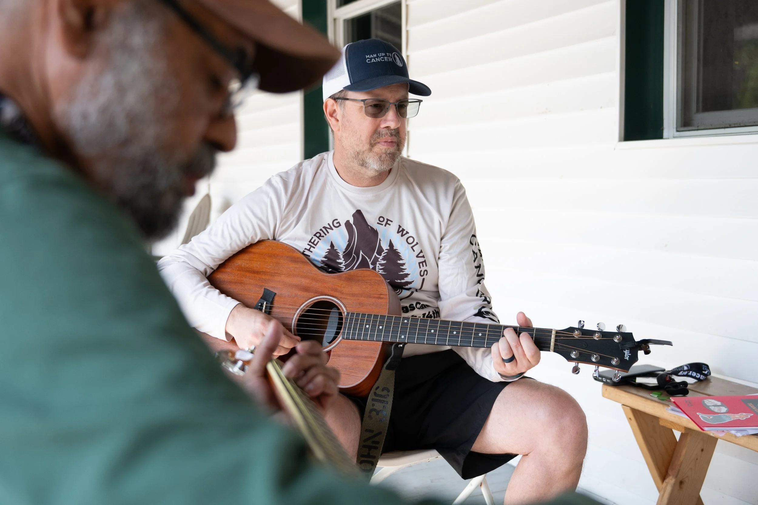 Two men playing guitars outdoors on a porch, one wearing a white long-sleeve shirt with a graphic design and a baseball cap, the other seen blurred in the foreground.