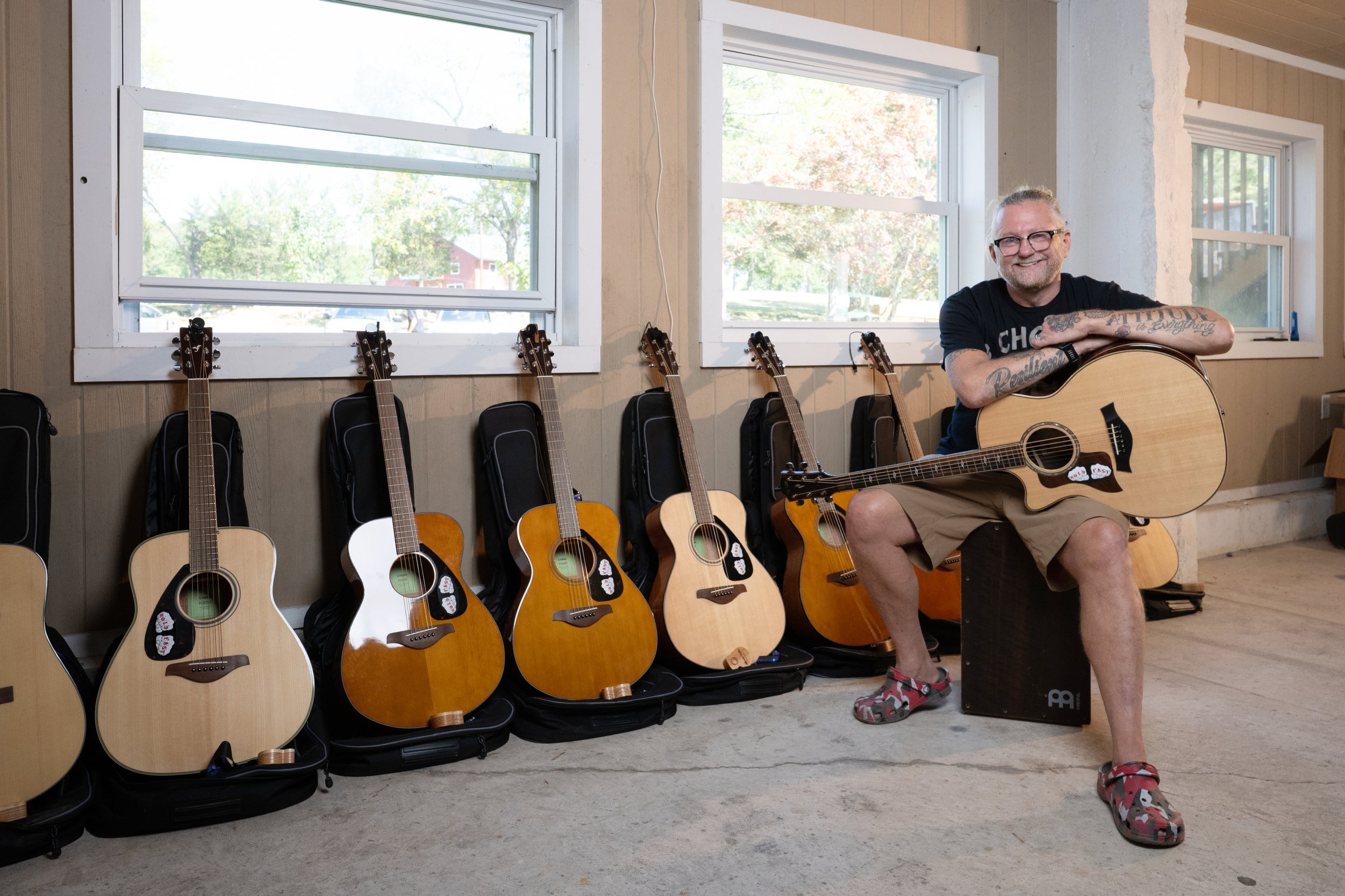 Man with glasses sitting on an acoustic speaker holding a guitar, with six more guitars on stands behind him.