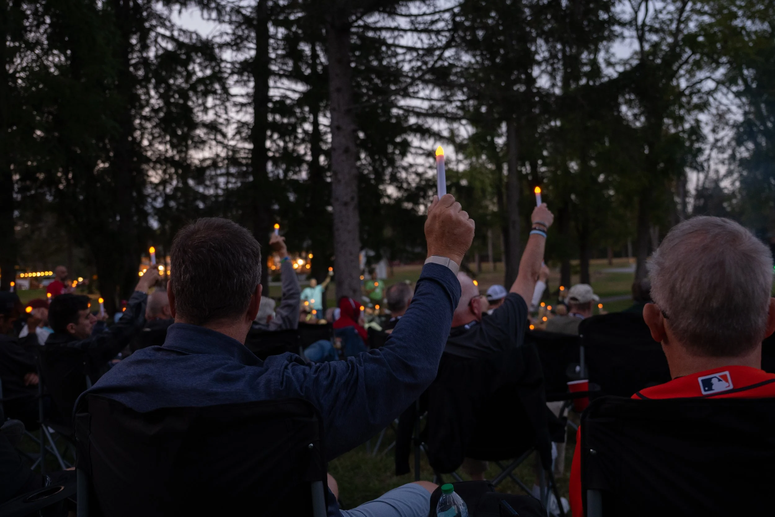 People gathered outdoors during evening, holding lit candles in a candlelight vigil, surrounded by tall trees.