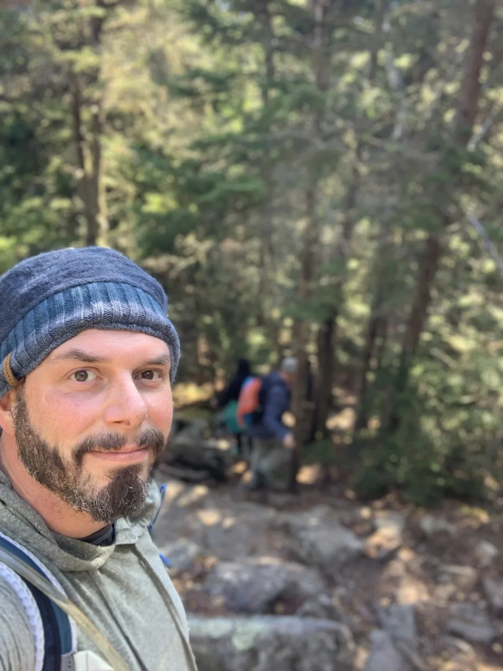 A man with a beard and a beanie taking a selfie on a forest trail, with two other hikers in the background among tall trees.