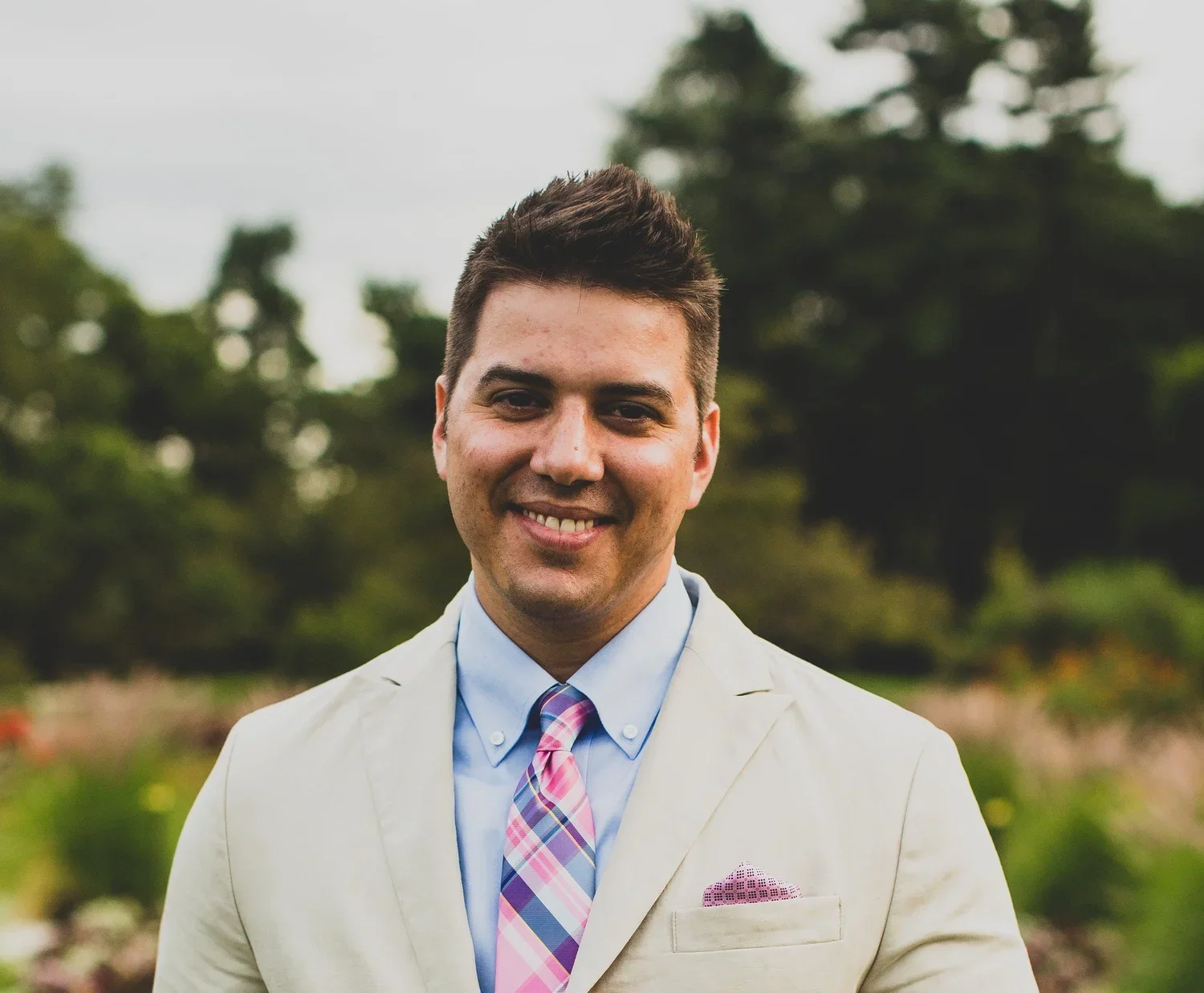 A man smiling outdoors, wearing a light-colored suit, blue shirt, and pink plaid tie, with a pink pocket square, standing in a green, tree-filled background.
