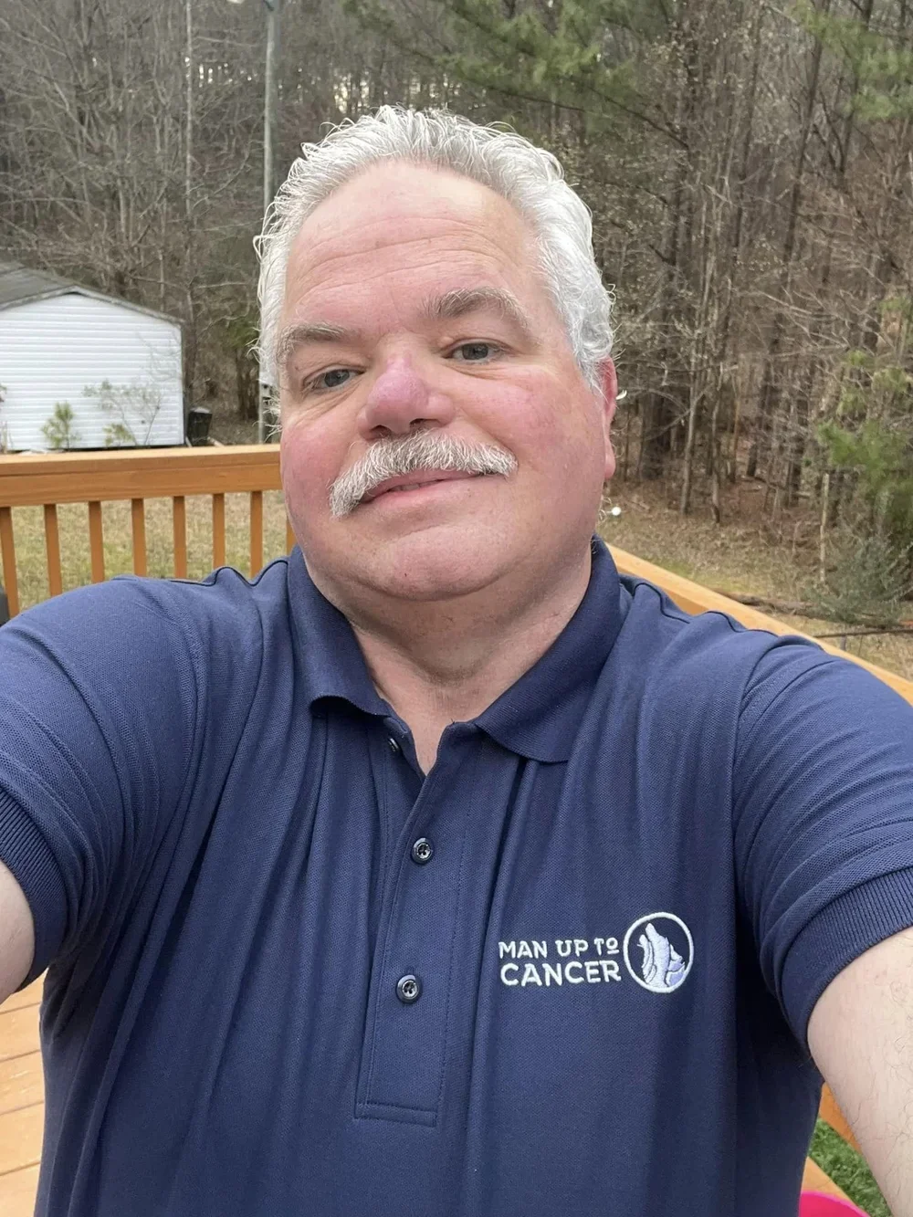 A man with gray hair and a mustache taking a selfie outdoors on a wooden deck. He is wearing a navy blue polo shirt with a logo and text that says 'Man Up To Cancer'. In the background, there are trees and a small white shed.