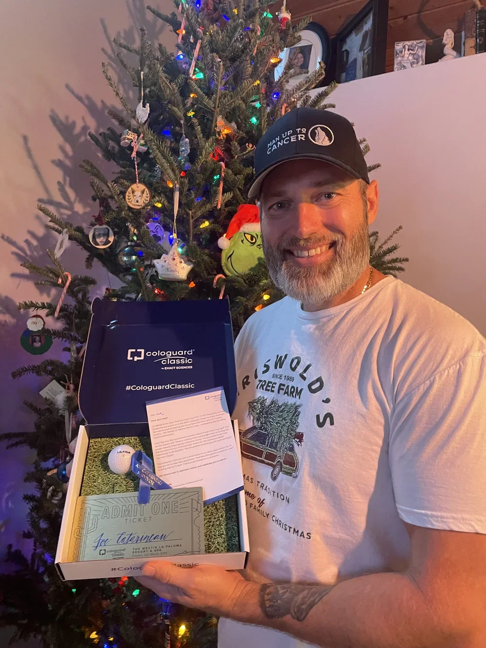 A man with a beard and a tattoo on his arm smiling and holding a box with a golf ball, a ticket, and a letter, standing in front of a decorated Christmas tree with colorful lights and ornaments.