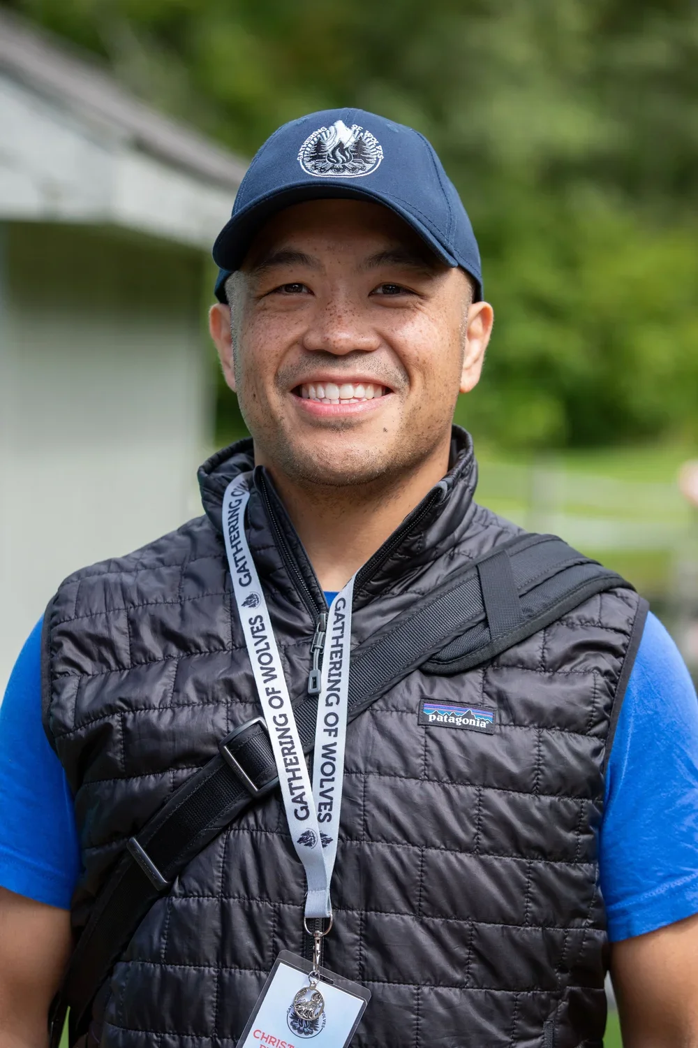 A smiling man outdoors wearing a blue Patagonia vest, a navy cap with a logo, a blue shirt, and a park ranger badge on a lanyard that says 'GATHERING OF WOLVES'.