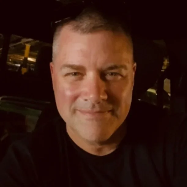 A smiling man with short hair taking a selfie indoors with a dark background.