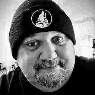 Close-up of a man wearing a black beanie with a logo of a howling wolf on it, smiling.