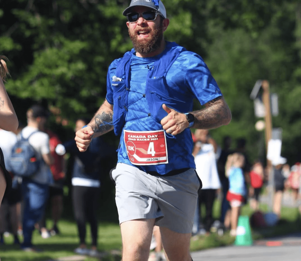 A man with a beard, sunglasses, and a blue athletic shirt running in a race, wearing a red bib number 4 at a Canada Day event.