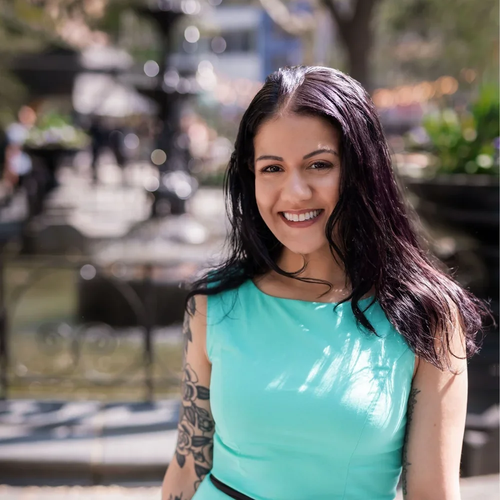 A woman smiling outdoors wearing a sleeveless turquoise top, with tattoos on her left arm and dark wavy hair, blurred background of trees and people in a park or urban setting.