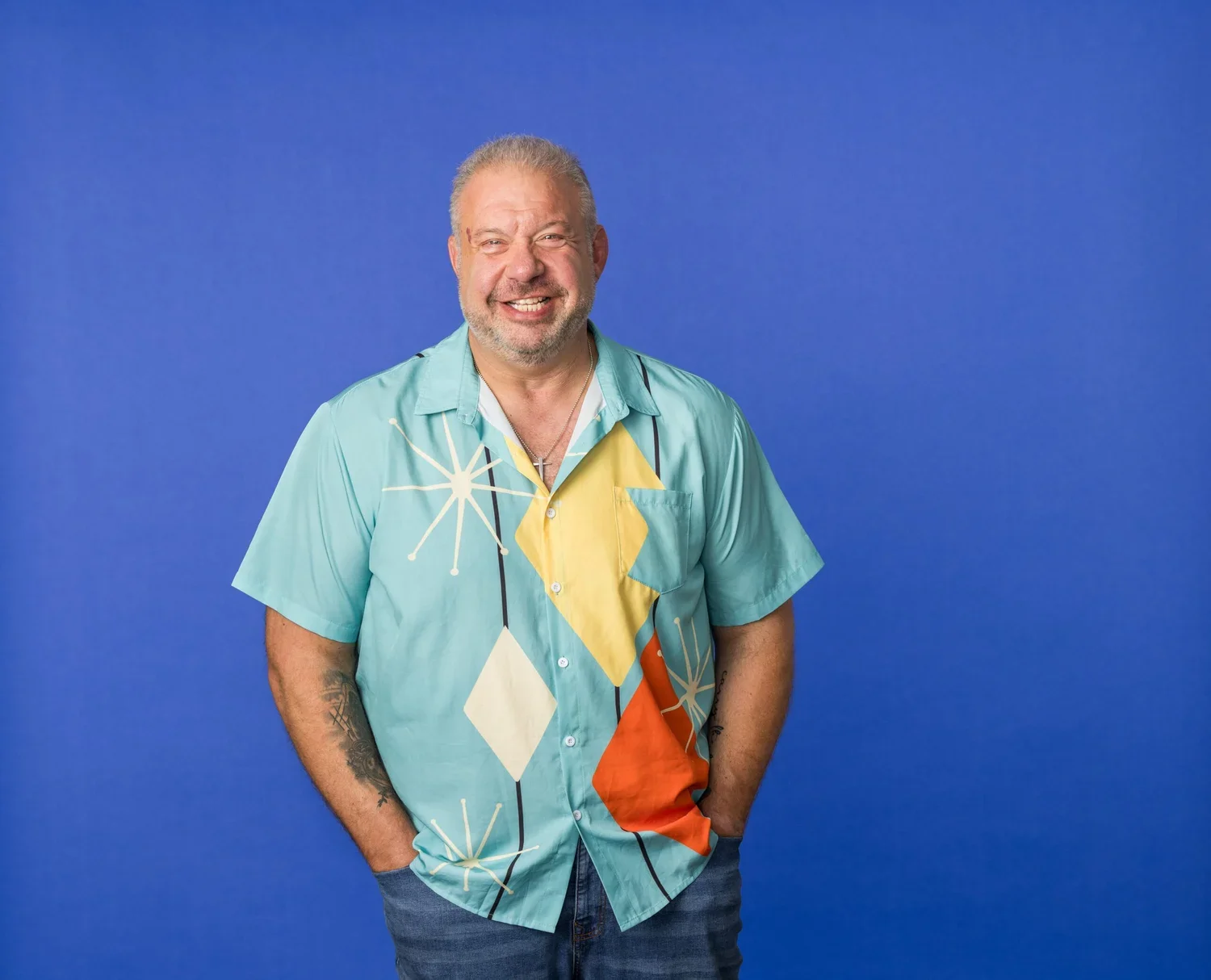 A smiling middle-aged man with a beard and tattoos, wearing a colorful, patterned short-sleeve shirt and jeans, standing against a solid blue background.