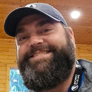 A smiling man with a beard wearing a gray cap indoors.