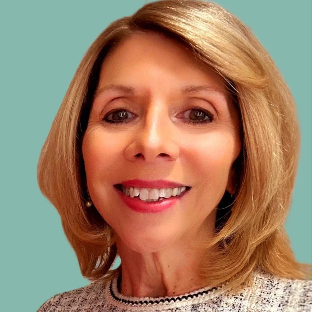 A woman with shoulder-length light brown hair, wearing a beige sweater with a dark trim and earrings, smiling against a teal background.