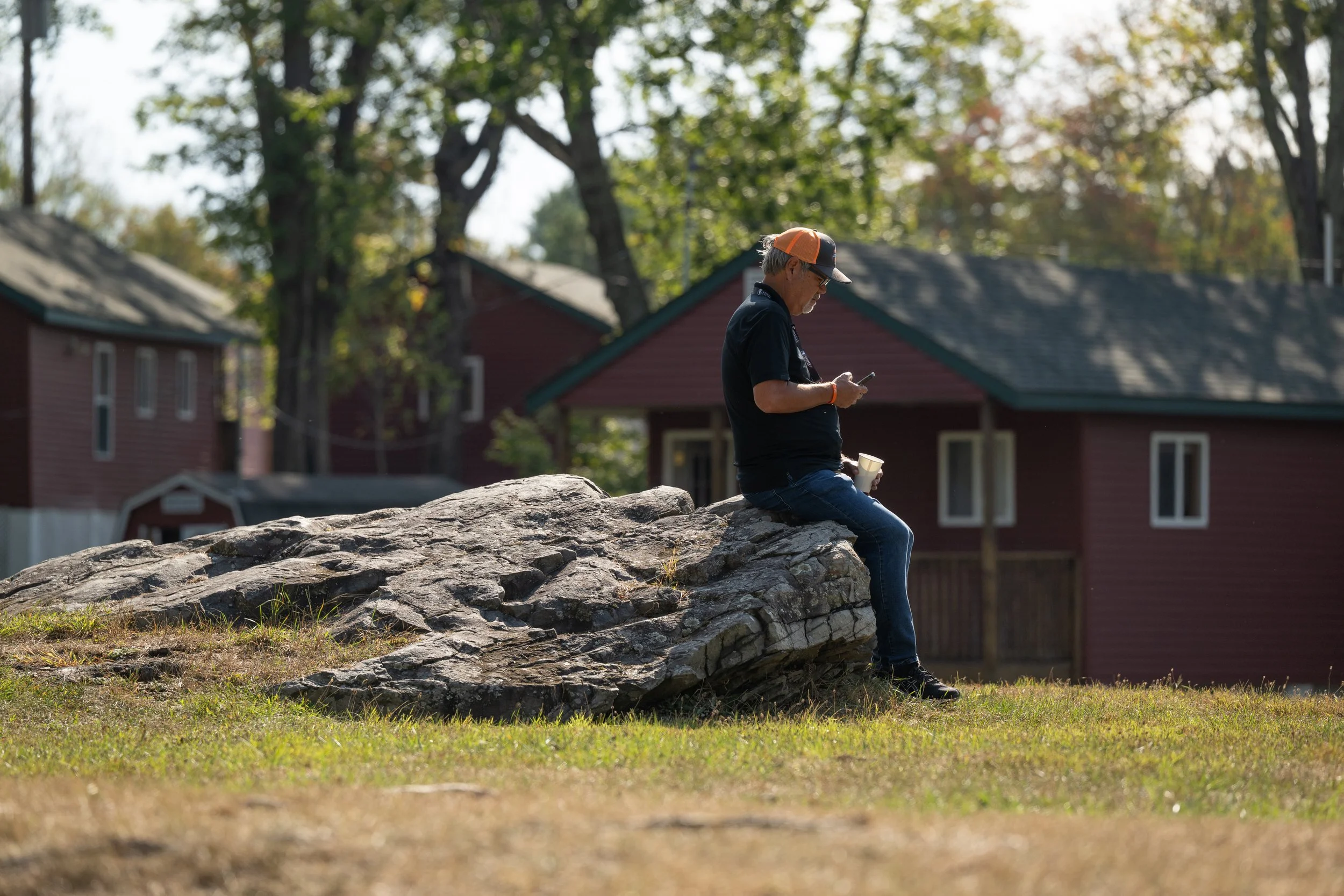 A man wearing a cap and glasses sitting on a rock, looking at his phone, with houses and trees in the background during the day.