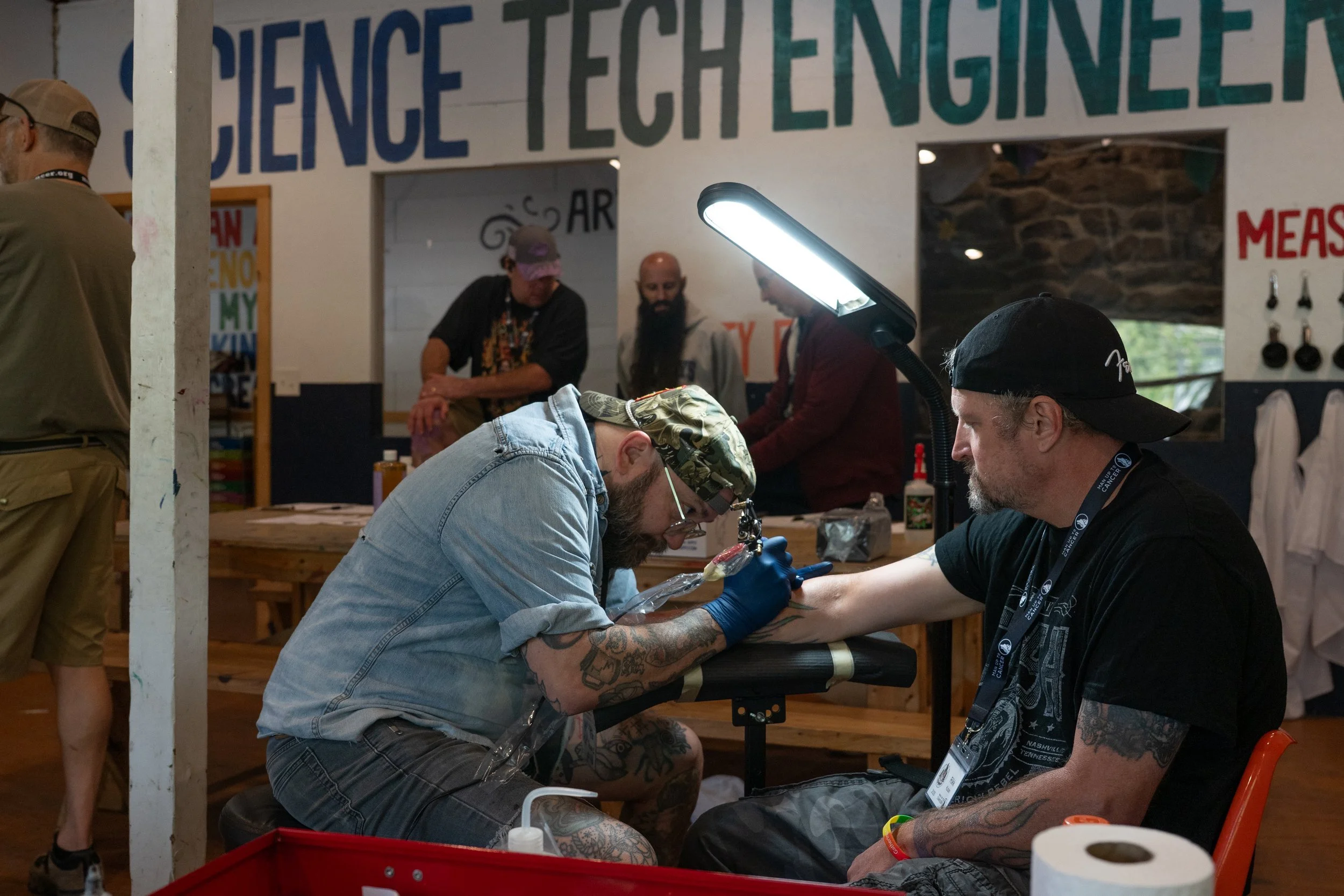 A man receives a tattoo on his arm from an artist in a tattoo shop, with several people in the background and a sign reading 'SCIENCE & TECHNOLOGY' on the wall.