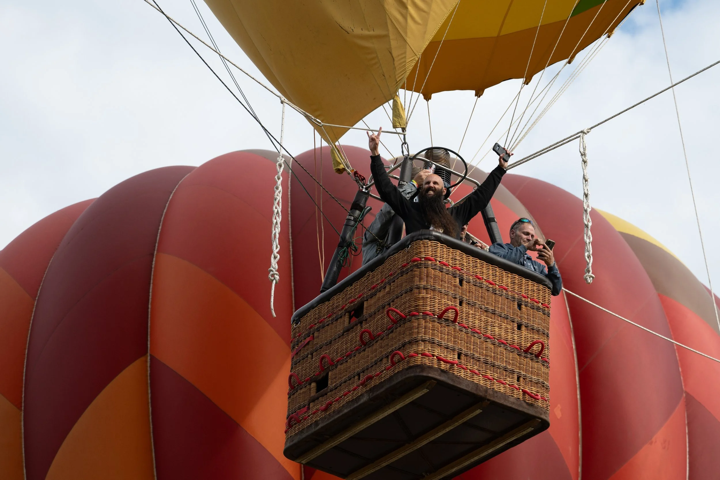 A hot air balloon basket with three passengers, two of whom are taking photos, against a backdrop of the envelope of the hot air balloon and the sky.