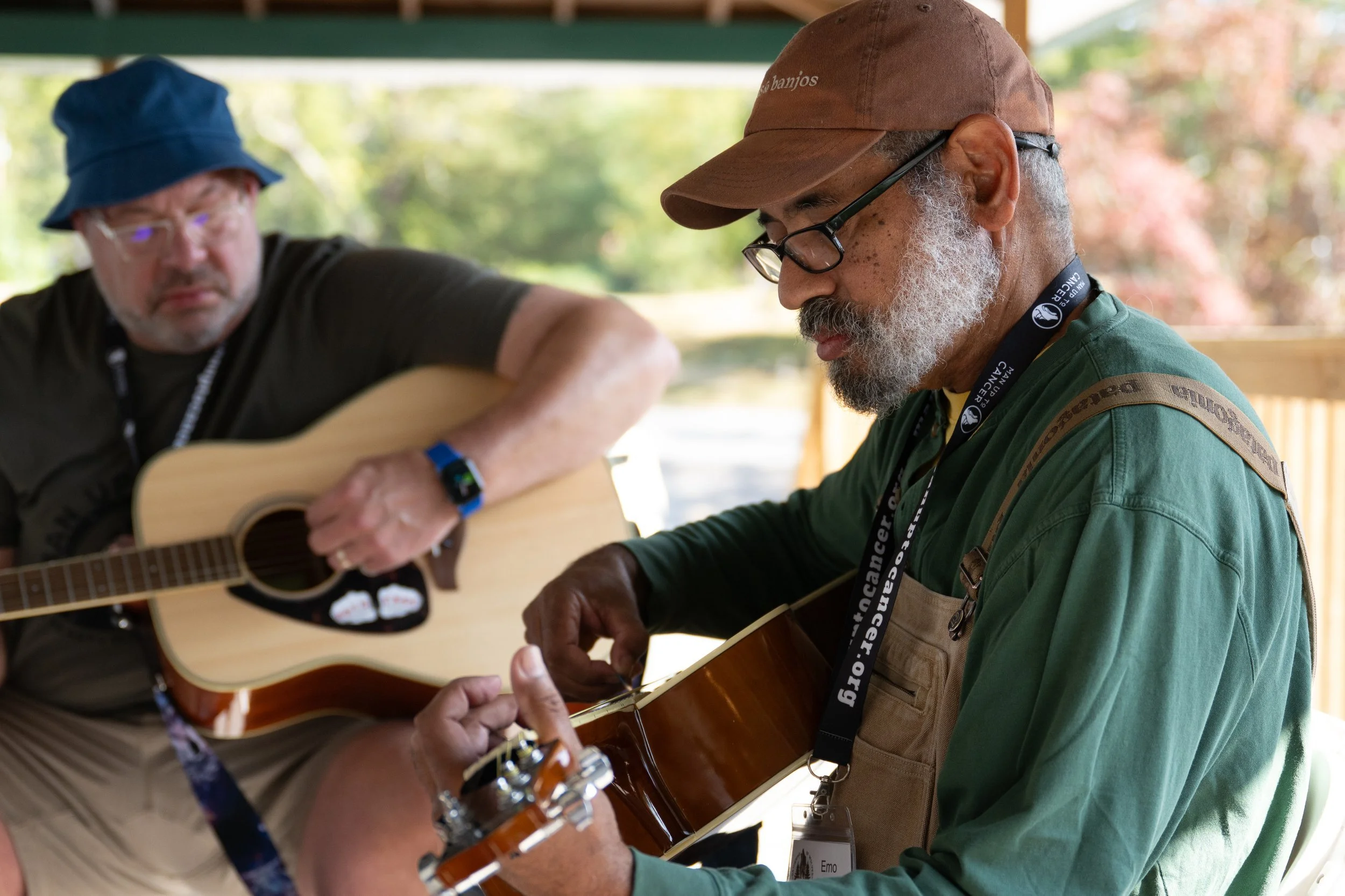Two men playing guitars, one with a blue hat and the other with a baseball cap, outdoors with blurred trees in the background.