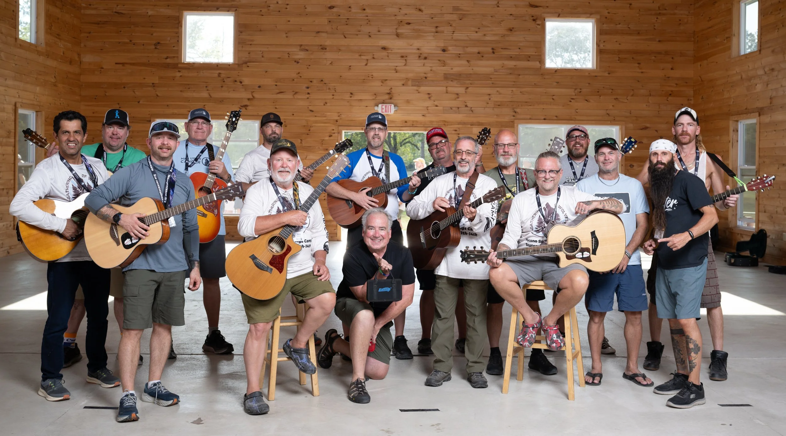Group of musicians with guitars inside a wooden hall, some sitting and others standing, smiling for a photo.