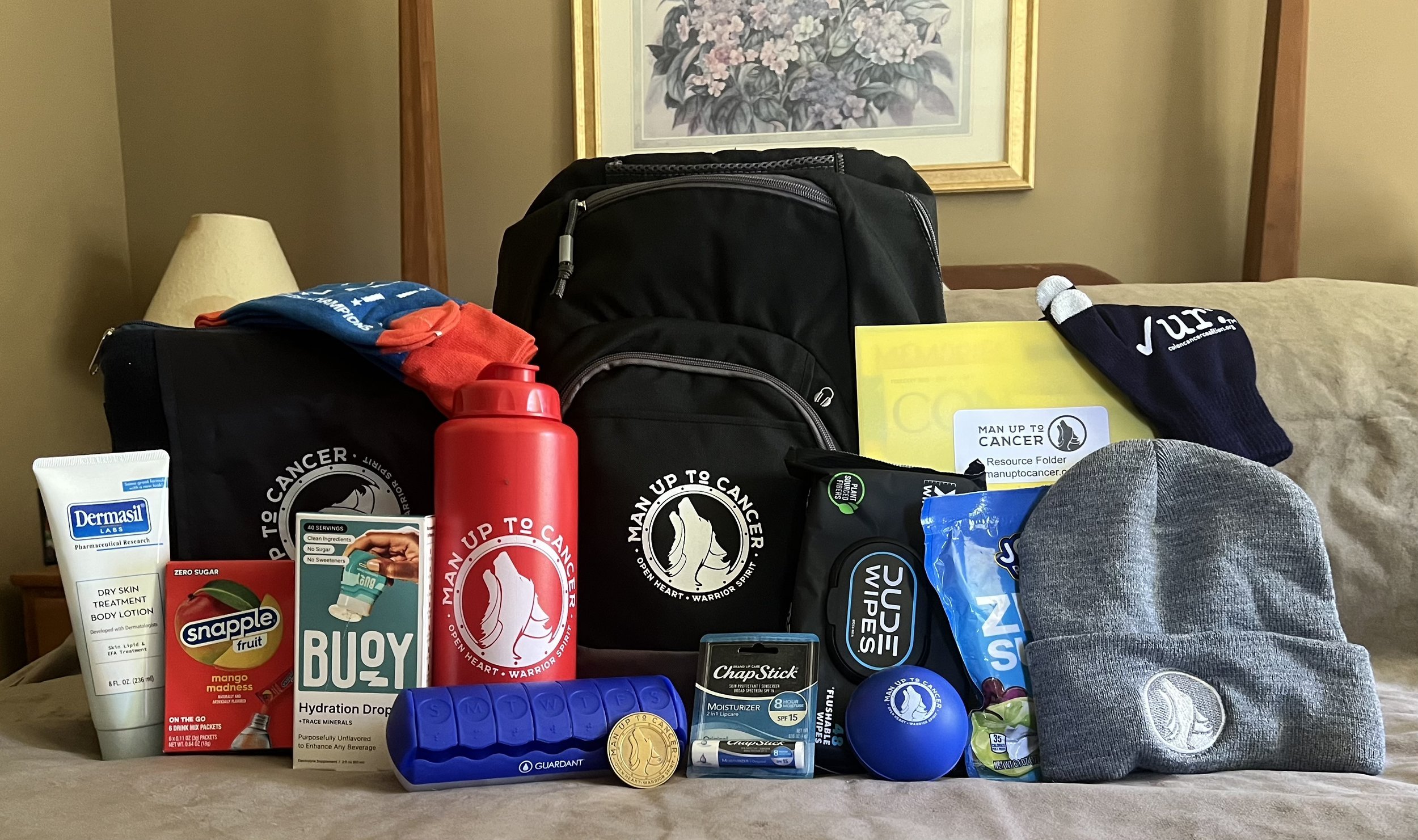 A collection of health and wellness supplies including medication, snacks, a backpack, a water bottle, a gray beanie, socks, and promotional materials related to cancer awareness, arranged in front of a framed floral picture on a beige bed.