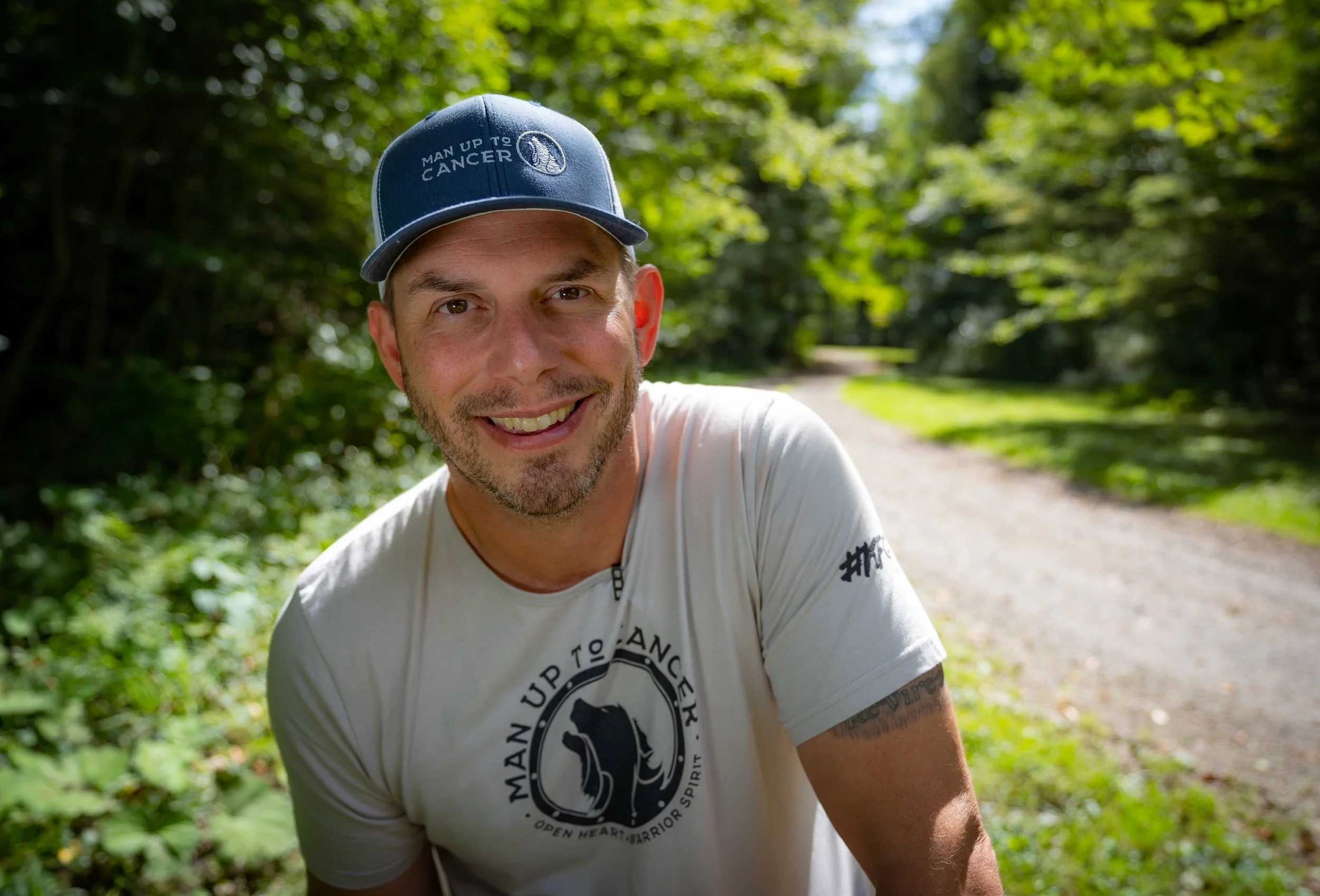 A smiling man wearing a blue baseball cap with the words "MAN UP TO CANCER" and a logo, standing outdoors in a wooded area with a dirt path behind him.
