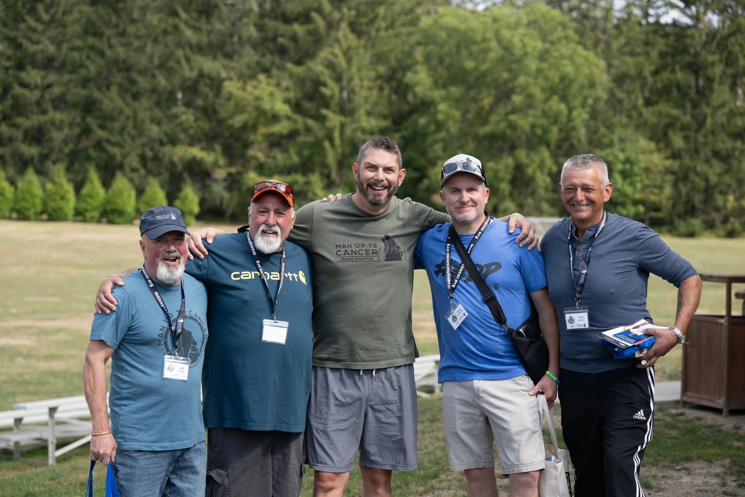 Five men with arms around each other smiling outdoors at a Man Up to Cancer event, wearing MUTC shirts and event lanyards.