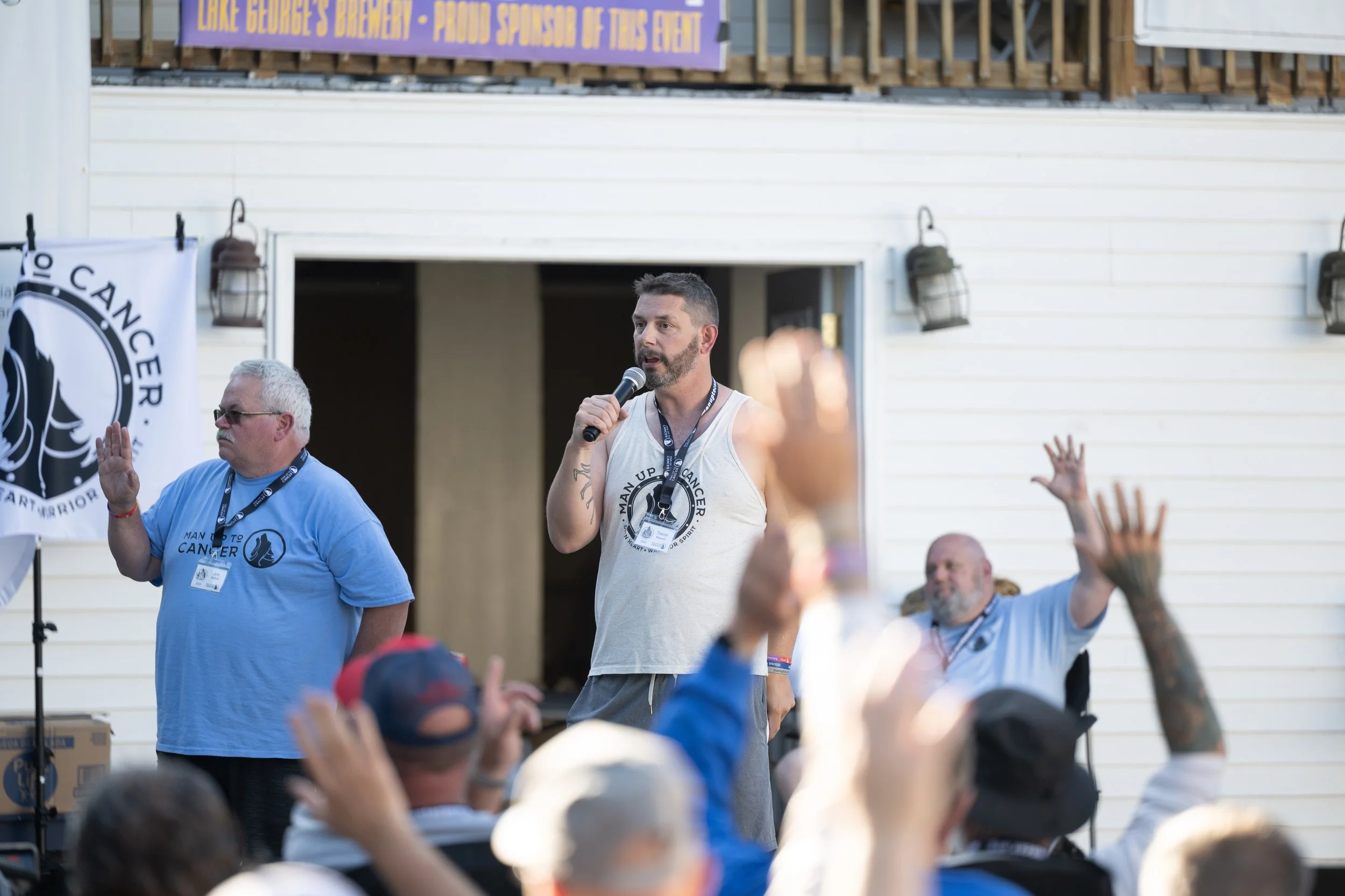Man in a Man Up to Cancer tank top speaking into a microphone at an outdoor event, with audience hands raised in the foreground.