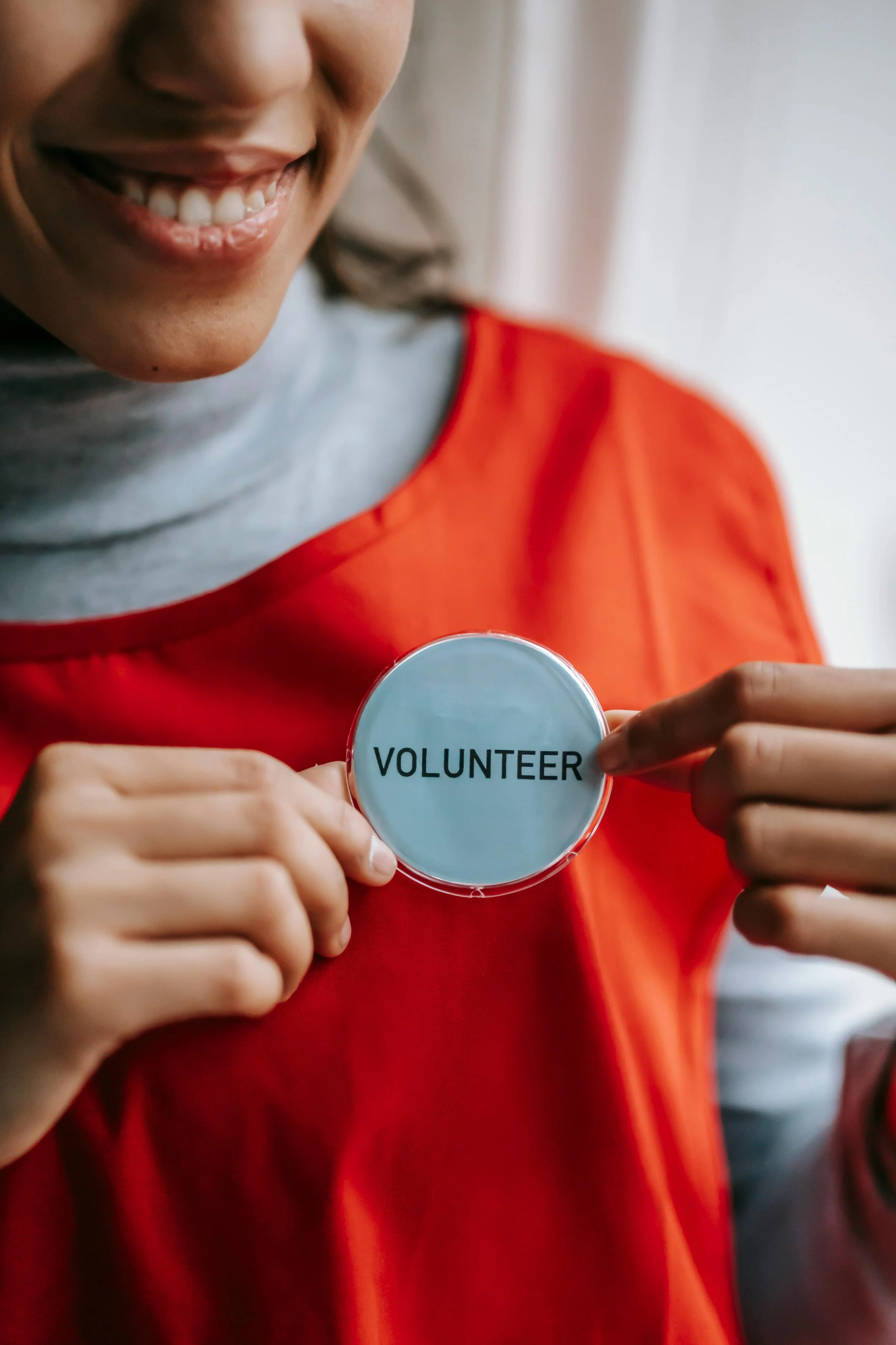 Smiling person in a red top holding a round "Volunteer" badge to their chest.