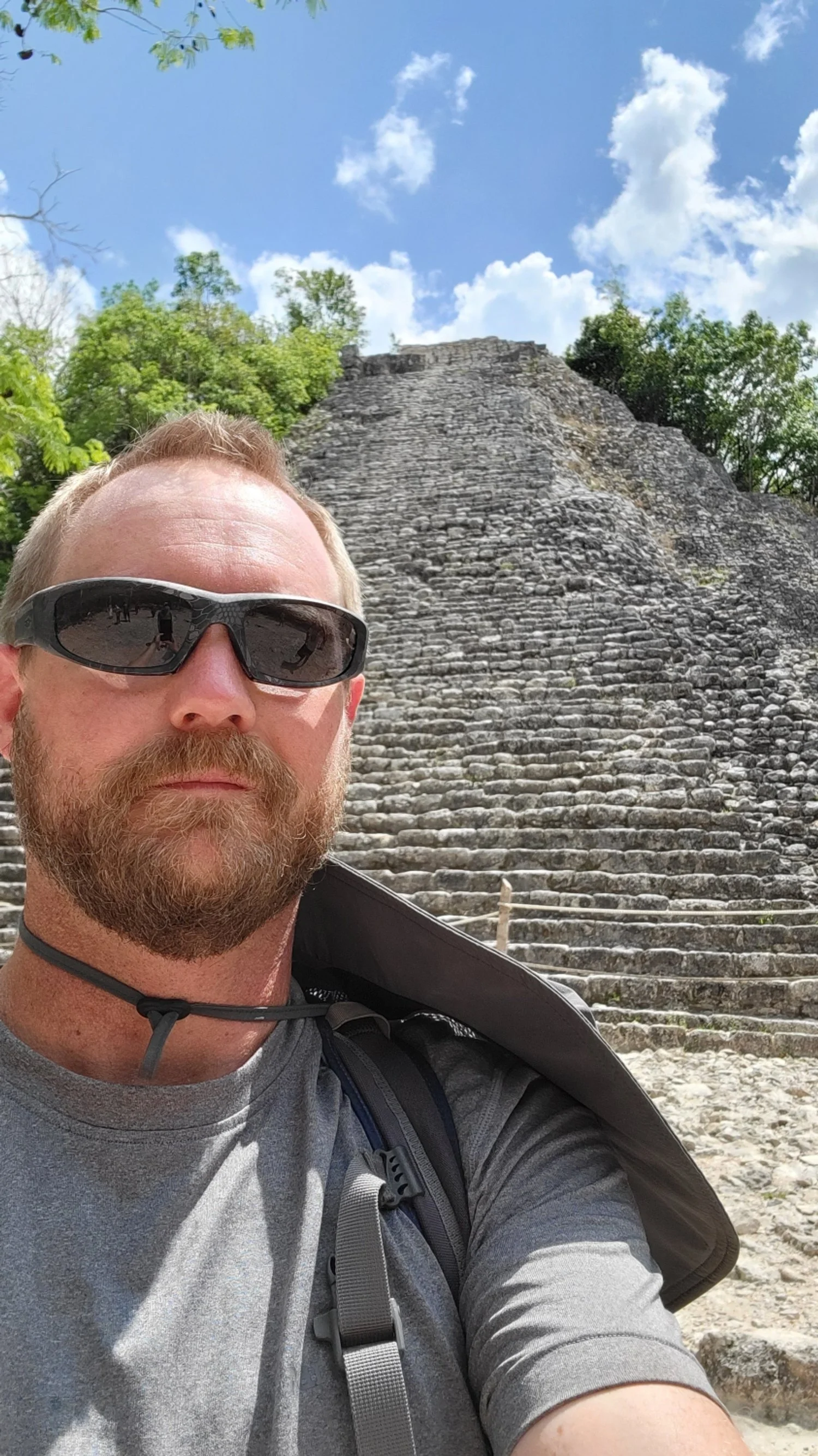A man with a beard and mustache, wearing a plaid shirt, looks thoughtfully to the side. A leafless tree is behind him against a clear blue sky. He is Co-Owner of Craving Amazing Travel. 