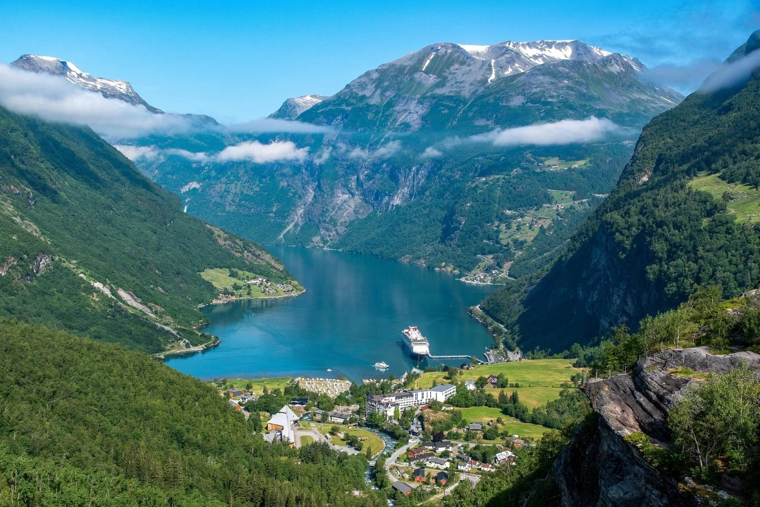 Scenic view of a fjord with a cruise ship docked near the small town at the base of green, rugged mountains with snow-capped peaks and scattered clouds in the sky.