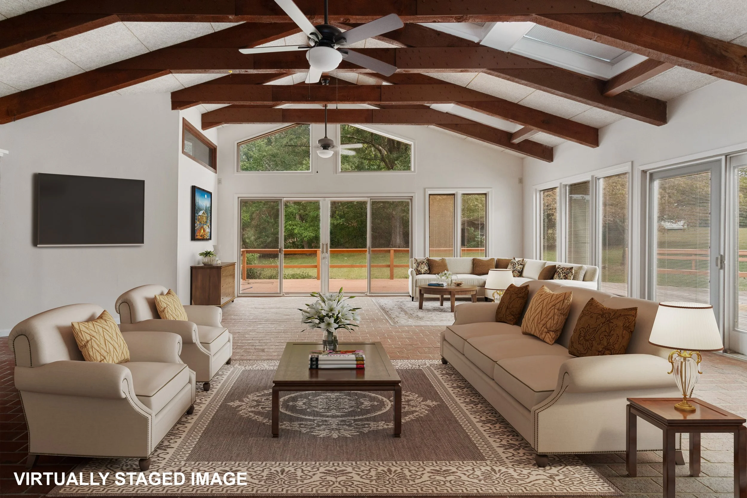 Spacious living room with vaulted ceiling, exposed wooden beams, and large windows overlooking greenery. Furniture includes a large sectional sofa, armchairs, a wooden coffee table with flowers, and side lamps. There's a TV mounted on the wall and a 