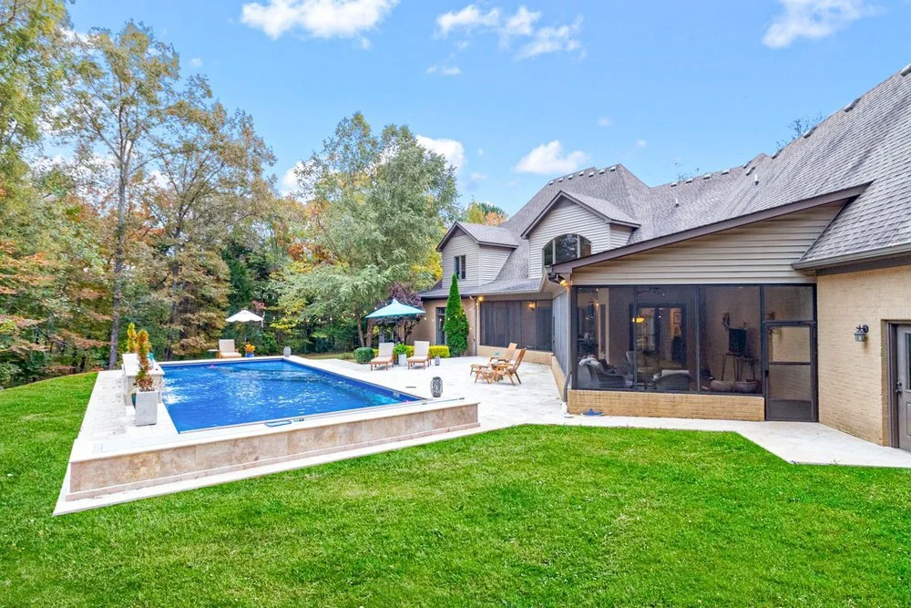Backyard with a swimming pool, lounge chairs, umbrellas, and patio furniture next to a house with a screened porch, surrounded by trees and green grass under a blue sky.
