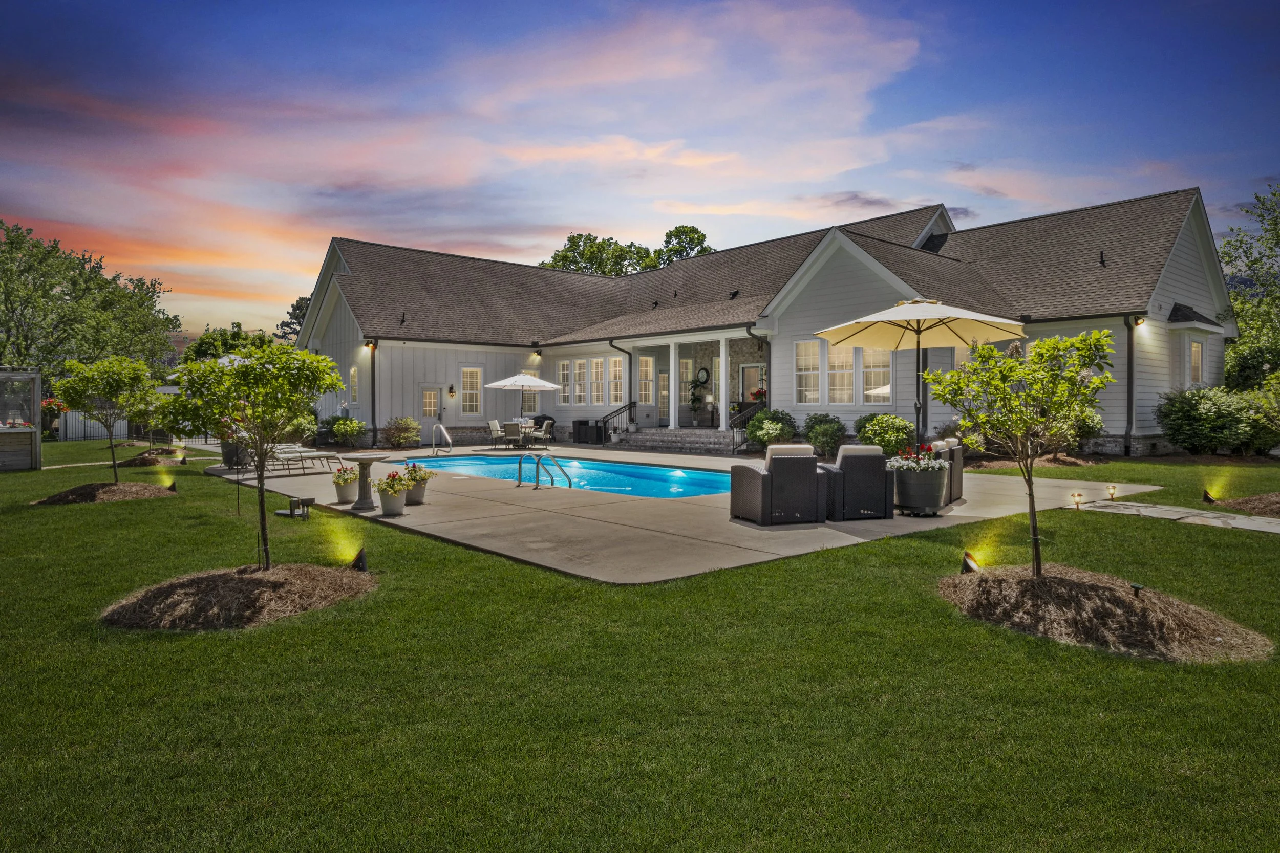 Backyard with a swimming pool, patio furniture, umbrellas, and surrounding trees at dusk.
