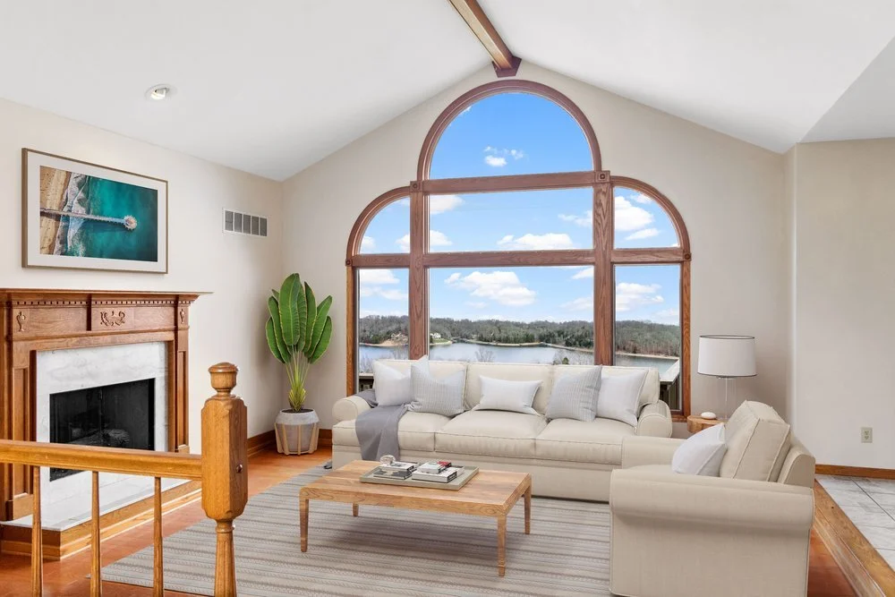 Living room with a large window showing a lakeside view, a white sofa with multiple cushions, a wooden coffee table, a fireplace with wooden mantel, a tall green plant, a floor lamp, and a framed ocean photograph on the wall.