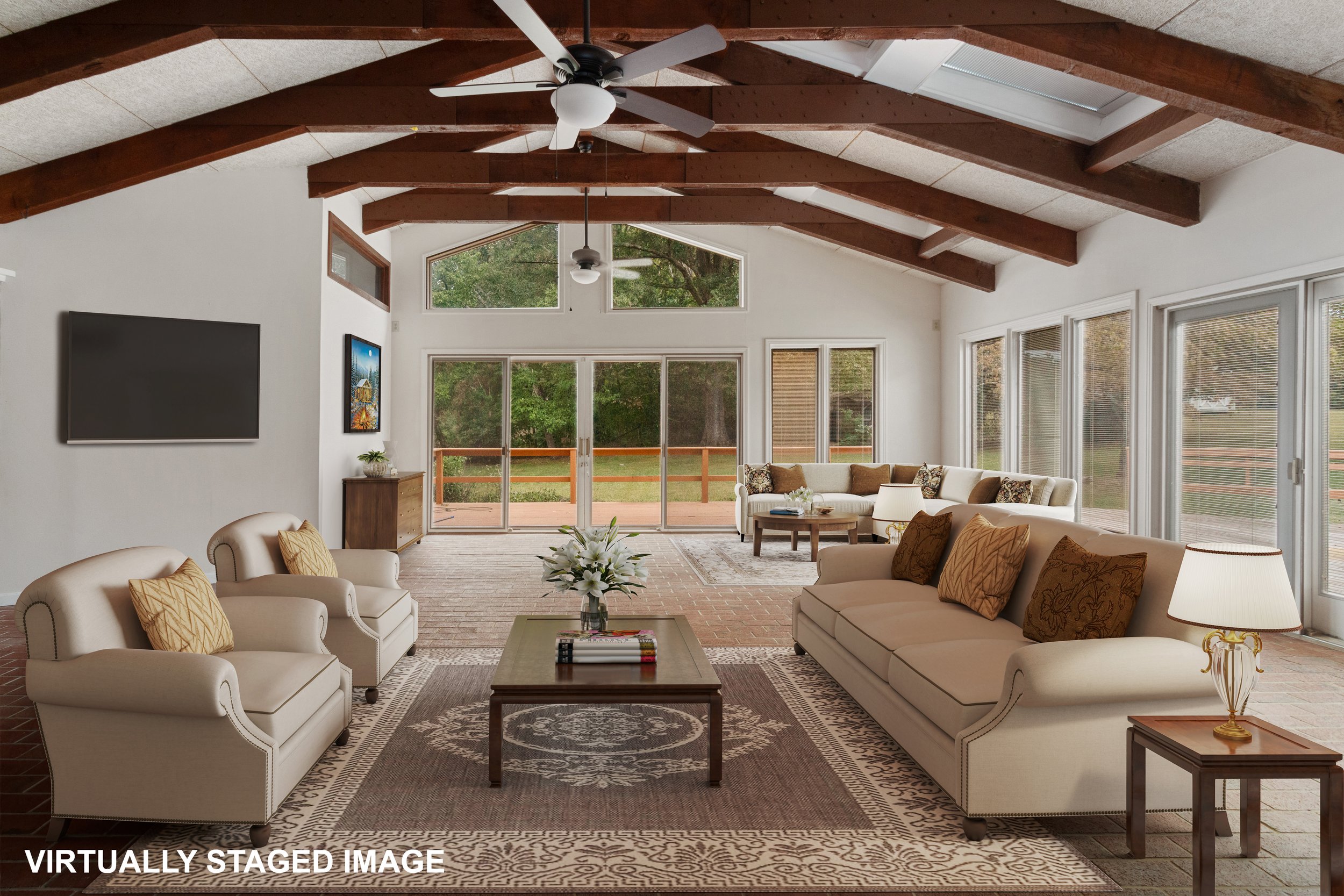 A spacious living room with high vaulted ceilings featuring exposed wooden beams, large windows, and sliding glass doors leading to an outdoor deck. The room is furnished with sofas and armchairs, a coffee table, and lamps, with a mounted flat-screen TV on the wall.