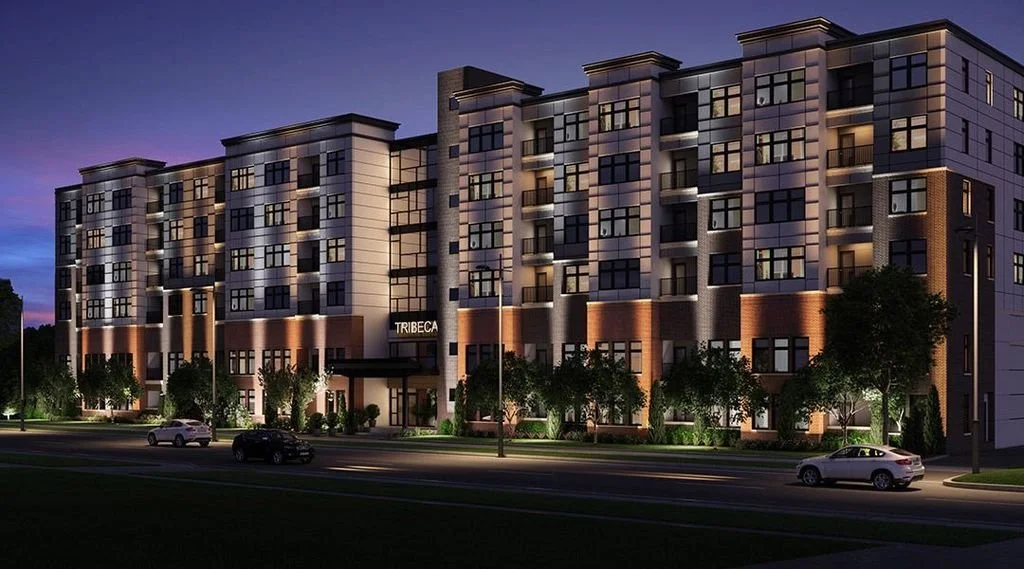 Night view of a modern apartment building with lit windows, trees along the sidewalk, and several cars on the street in front. The building has a sign that says 'TRIBECA' above the entrance.