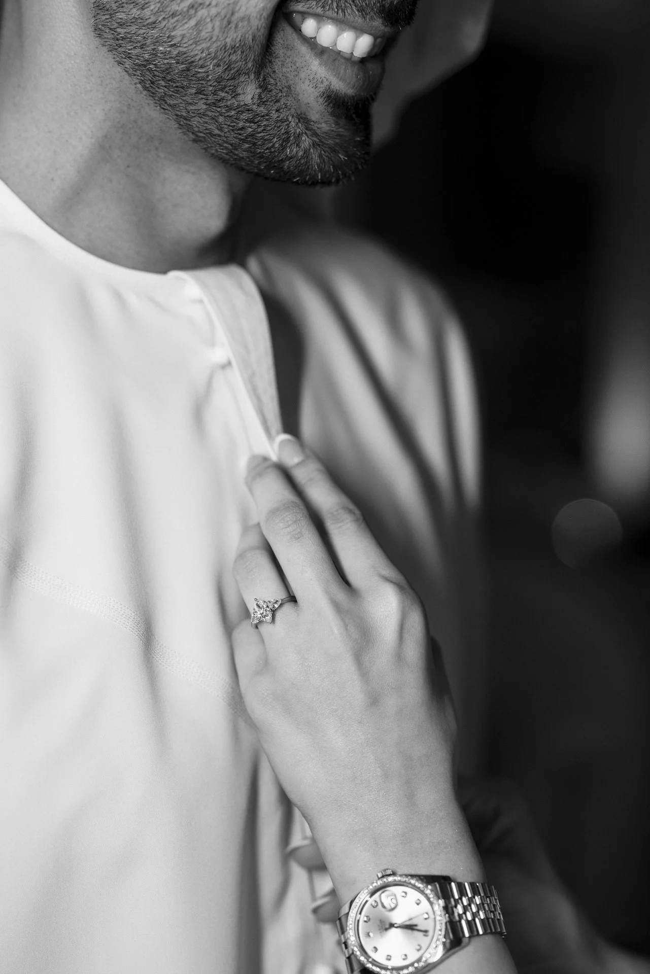 Close-up of a smiling man with a beard, wearing a watch and ring, adjusting his shirt collar with a woman’s hand resting on his chest.