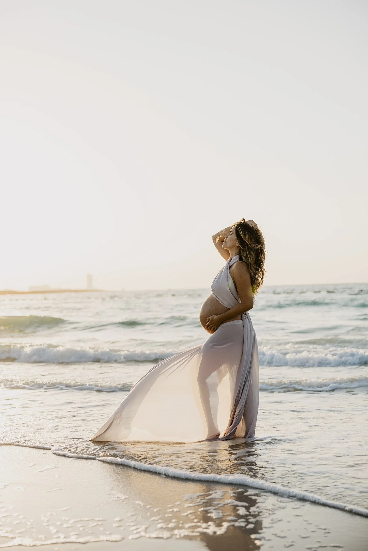 A pregnant woman wearing a flowing white dress standing at the edge of the ocean as waves wash around her feet, with a sunset or sunrise sky in the background.